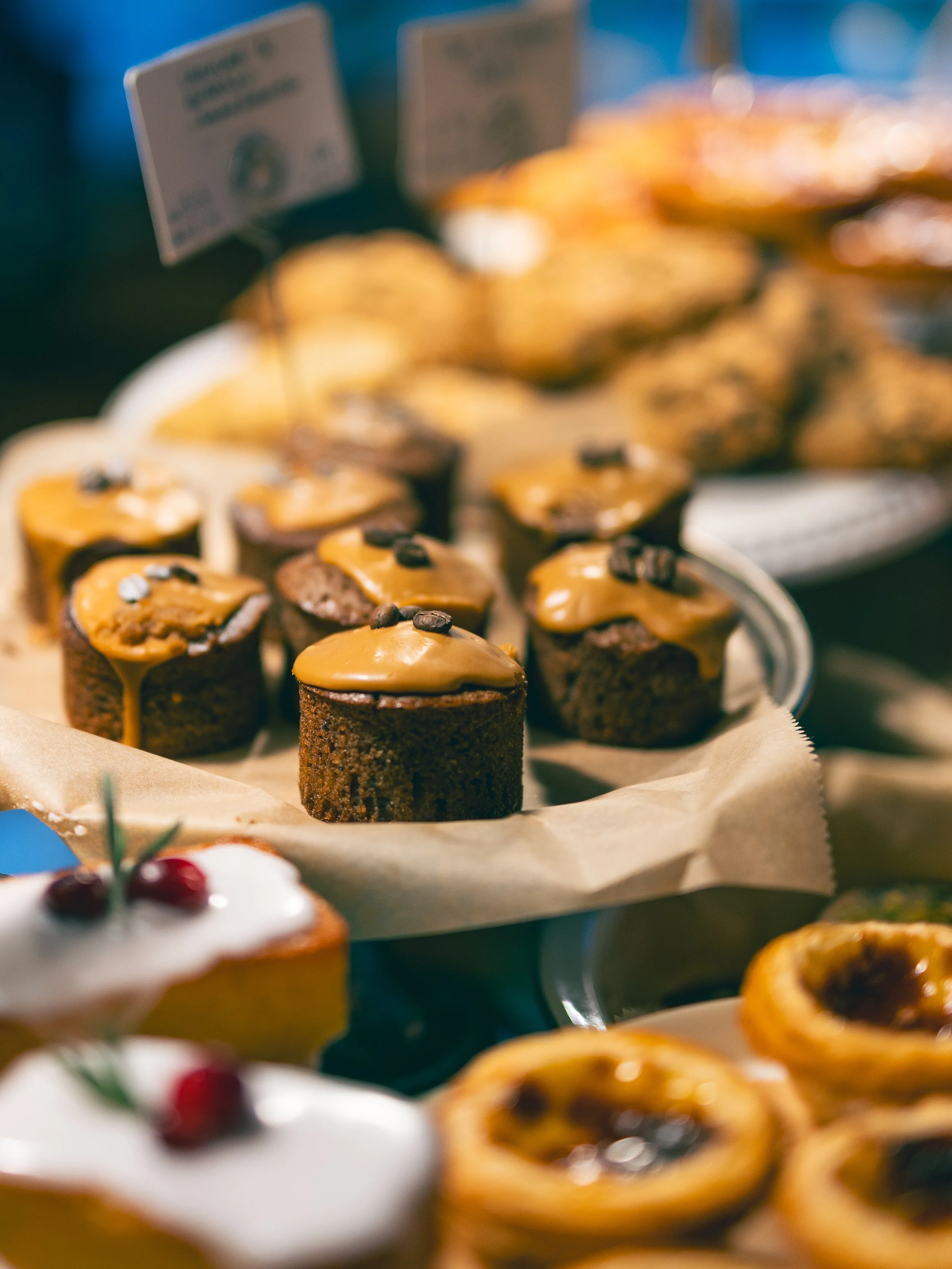 Assorted pastries on display at a bakery, including mini chocolate cupcakes with caramel frosting and coffee beans, and Portuguese egg tarts with a glossy surface.