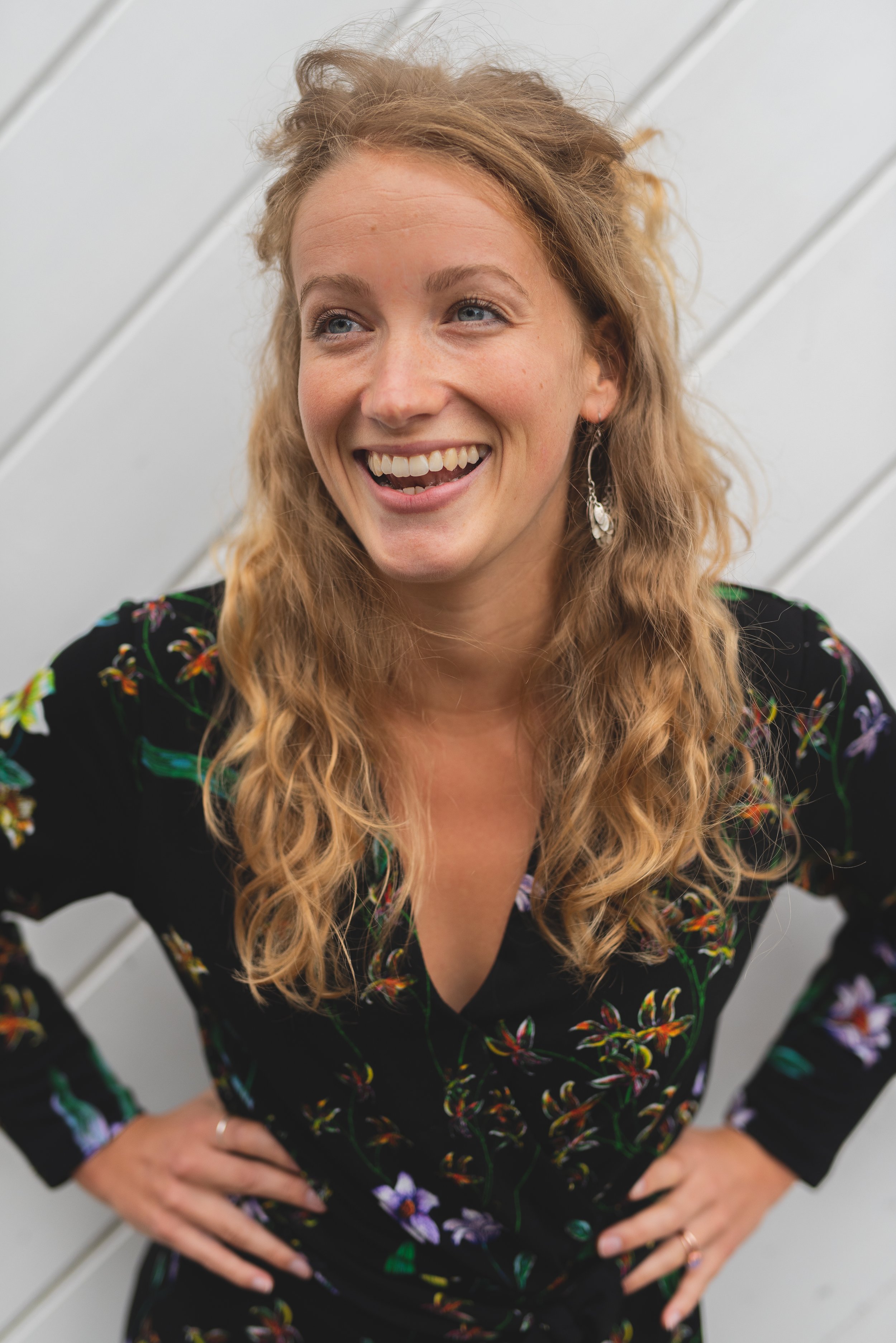 A woman with curly red hair smiling and looking to the side, standing against a white background.