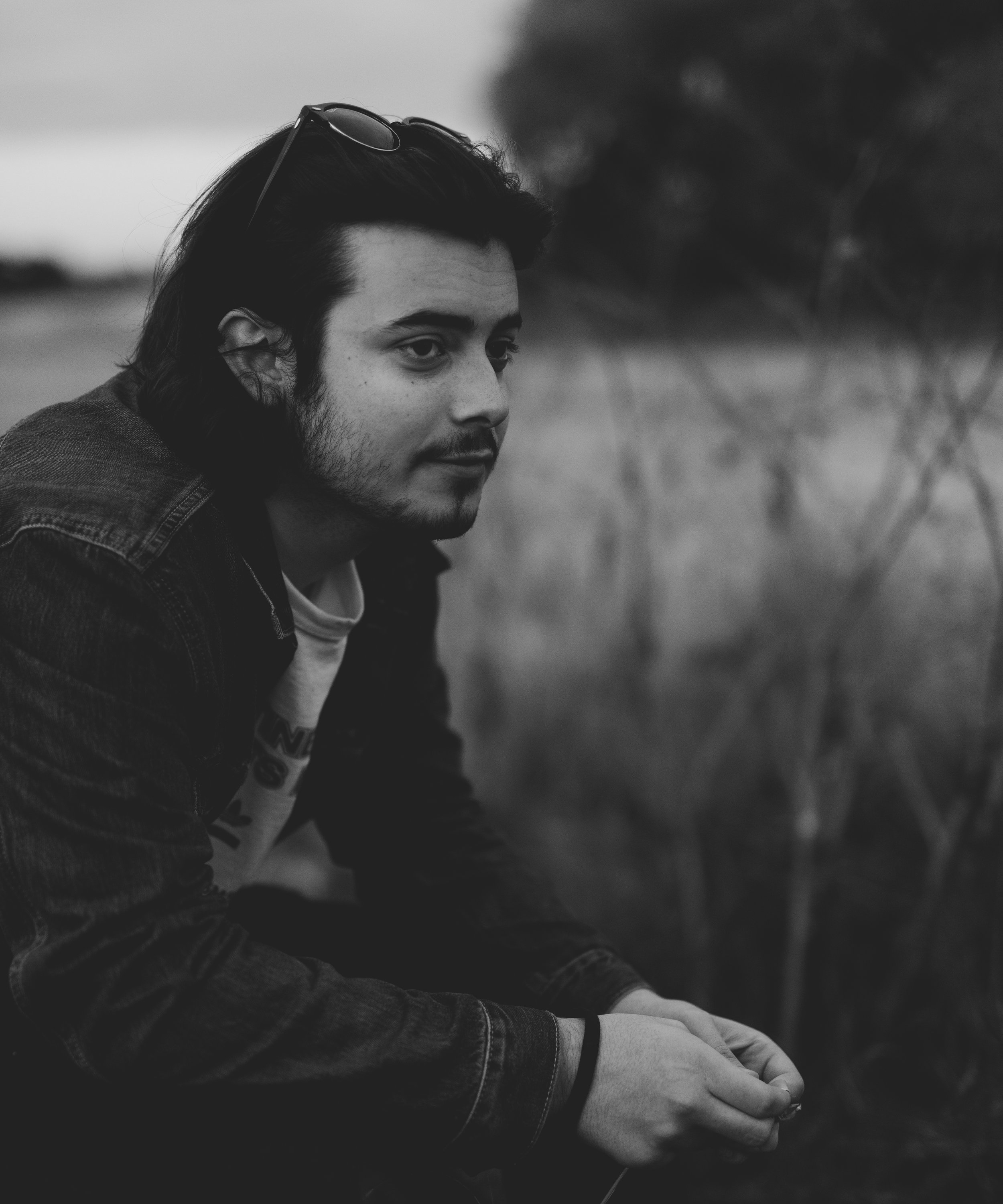 A young man with dark hair and beard, wearing a denim jacket and sunglasses on his head, crouches outdoors in a field with blurred trees in the background.
