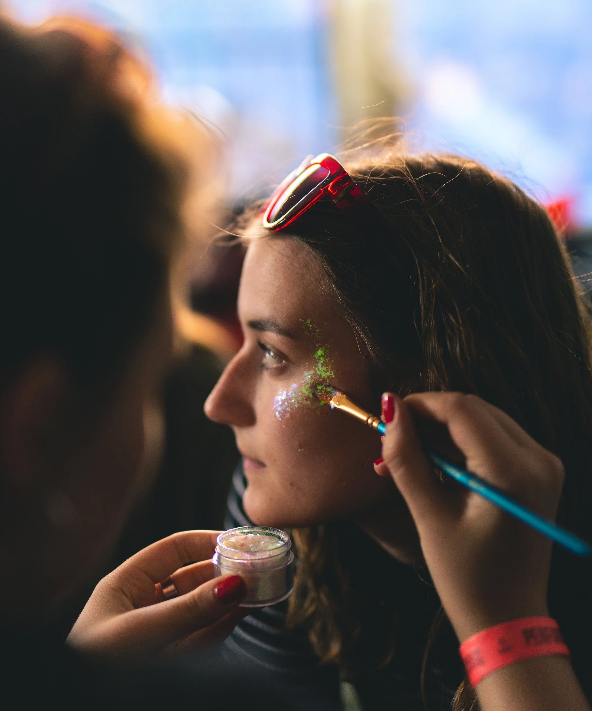 A woman with sunglasses on her head having glitter and face paint applied to her face by an artist at a festival or event.