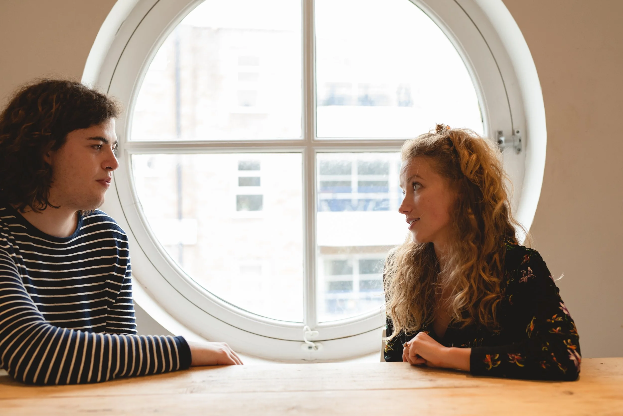 Two women sitting at a wooden table near a round window, engaged in a conversation. One woman has curly hair and wears a floral top, the other has dark hair and wears a striped shirt.
