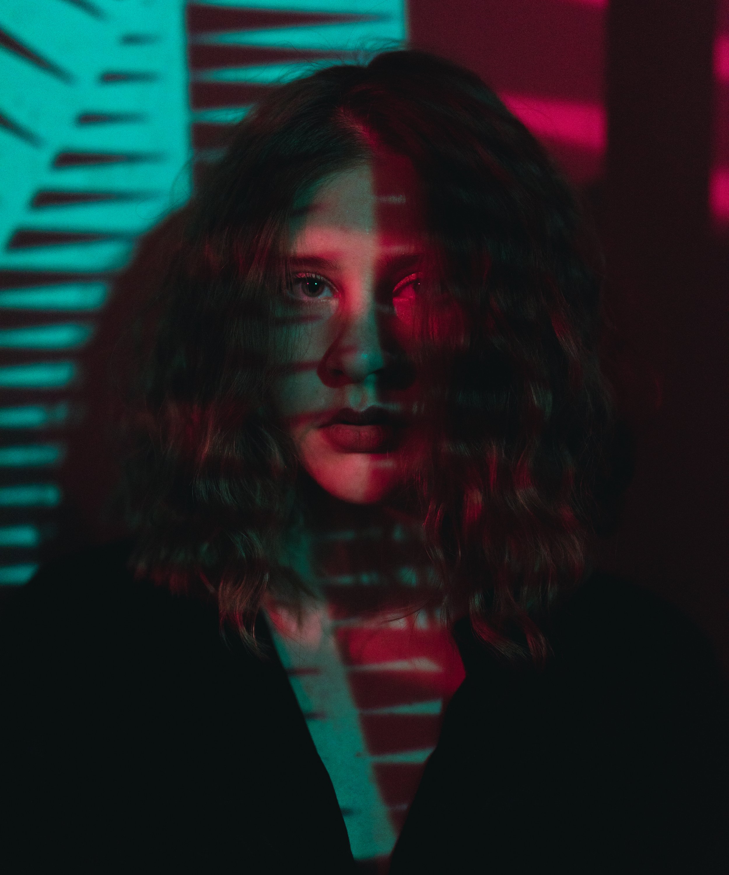 A woman with curly hair and bold makeup faces the camera with a neon light pattern casting shadows on her face, creating a moody atmosphere.