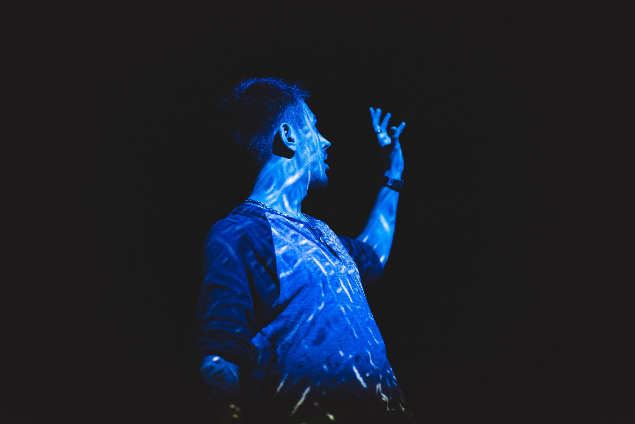 Side view of a young man with dark hair, wearing a tie-dye shirt, raising his right hand with fingers extended, against a dark background with blue lighting effects.