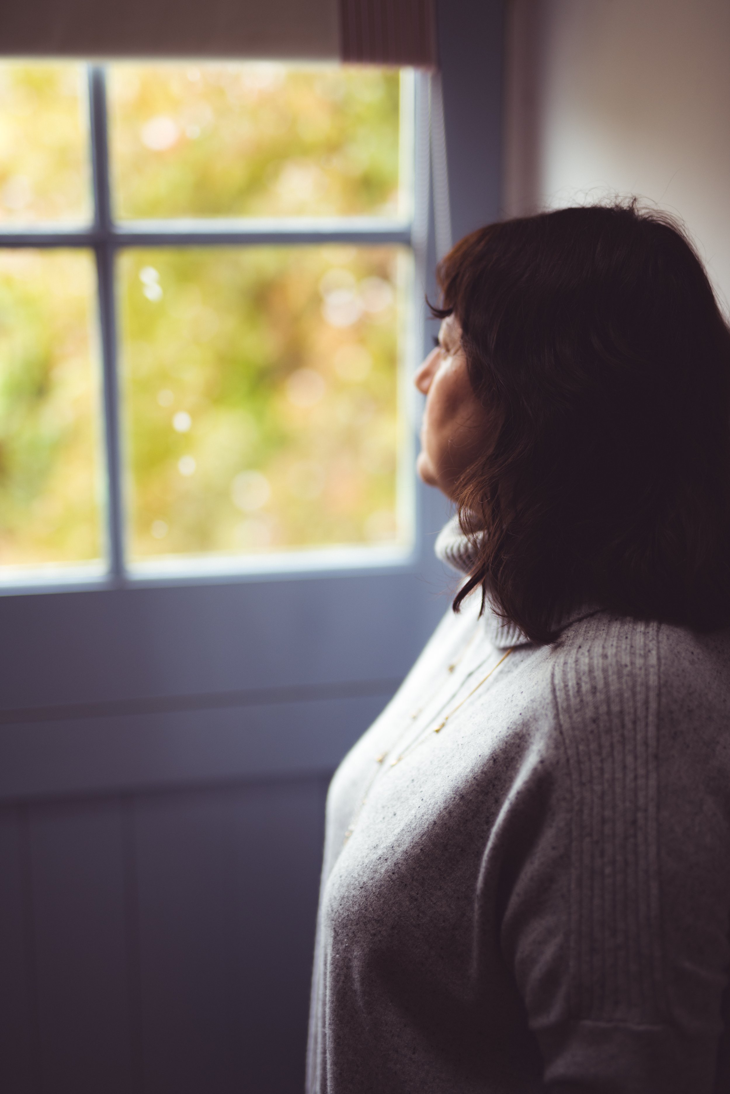 Woman with dark hair looking out a window during daytime, with blurred trees outside.