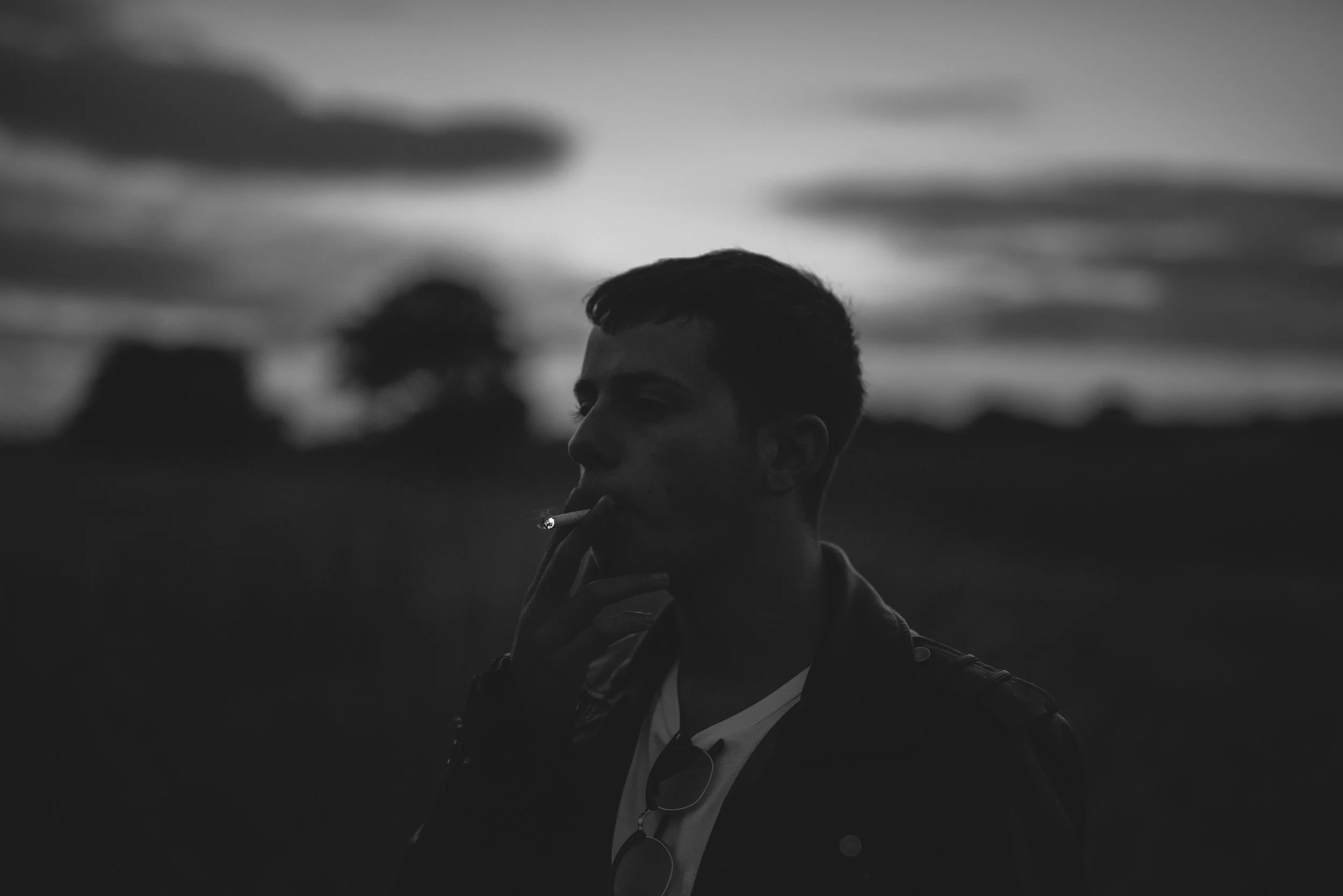 Silhouette of a young man smoking a cigarette during sunset or twilight, with a blurred background of trees and sky.