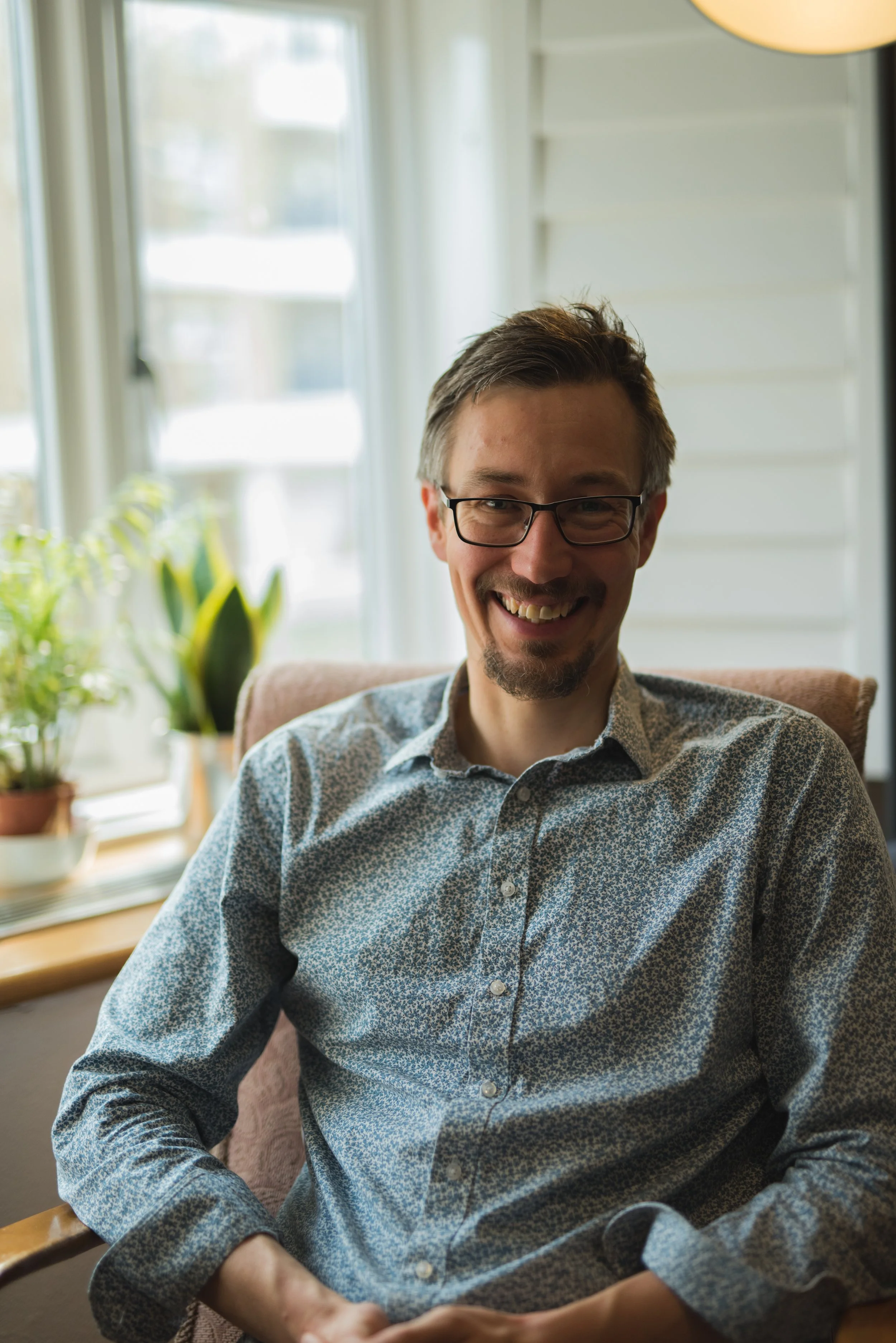 A smiling man with glasses and a patterned shirt sitting indoors with potted plants in the background.