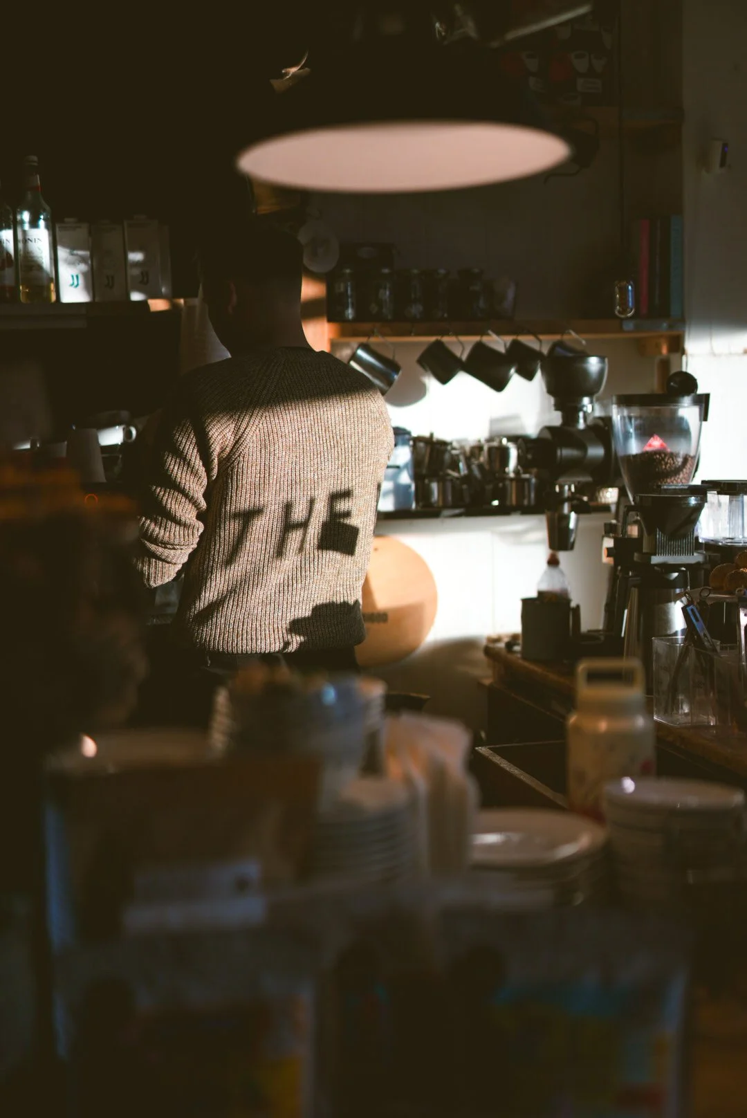 A person working behind a counter in a coffee shop or café, with coffee-making equipment and supplies visible around them.