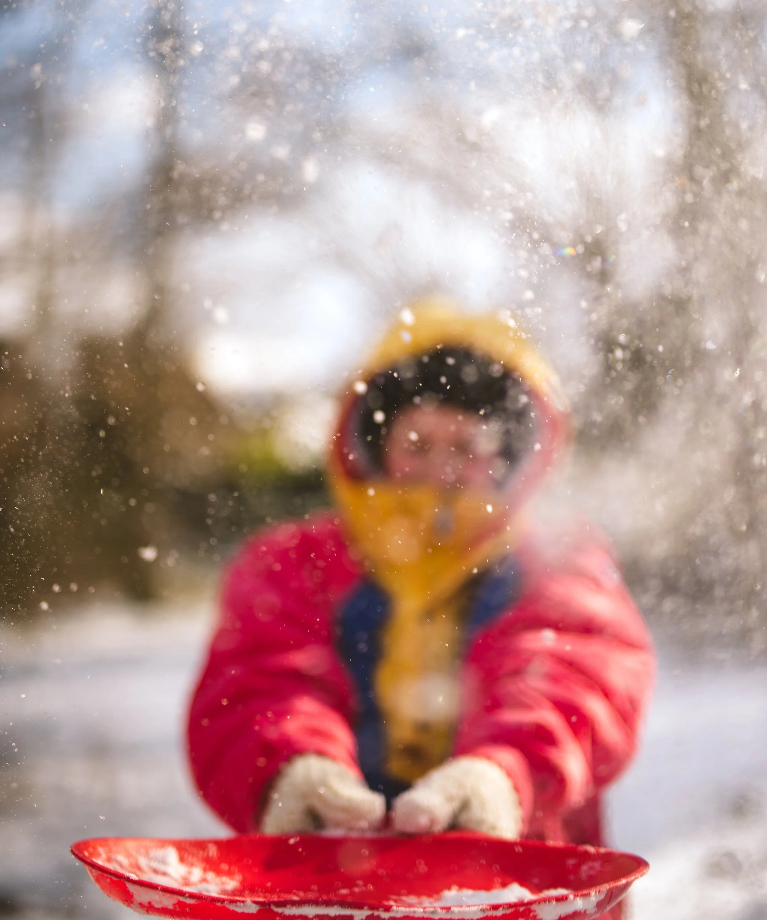 Child in a red jacket with gloves and a yellow hood holding a red sled in motion, with snow flying around during winter.