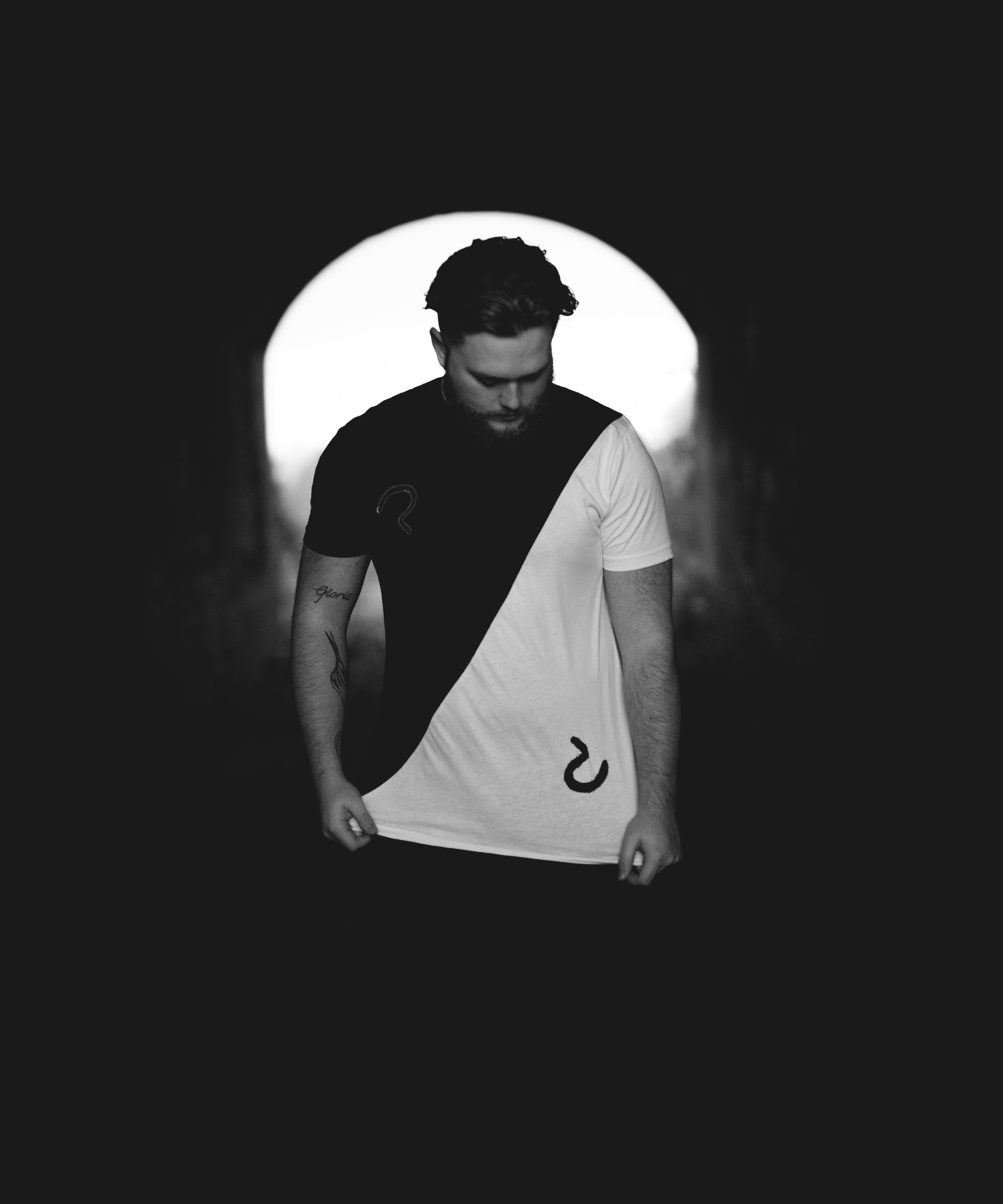 A young man with a beard is standing in a dark tunnel, with a bright light behind him. He is looking down and holding the bottom of his shirt, which has a logo on it divided into two colors.