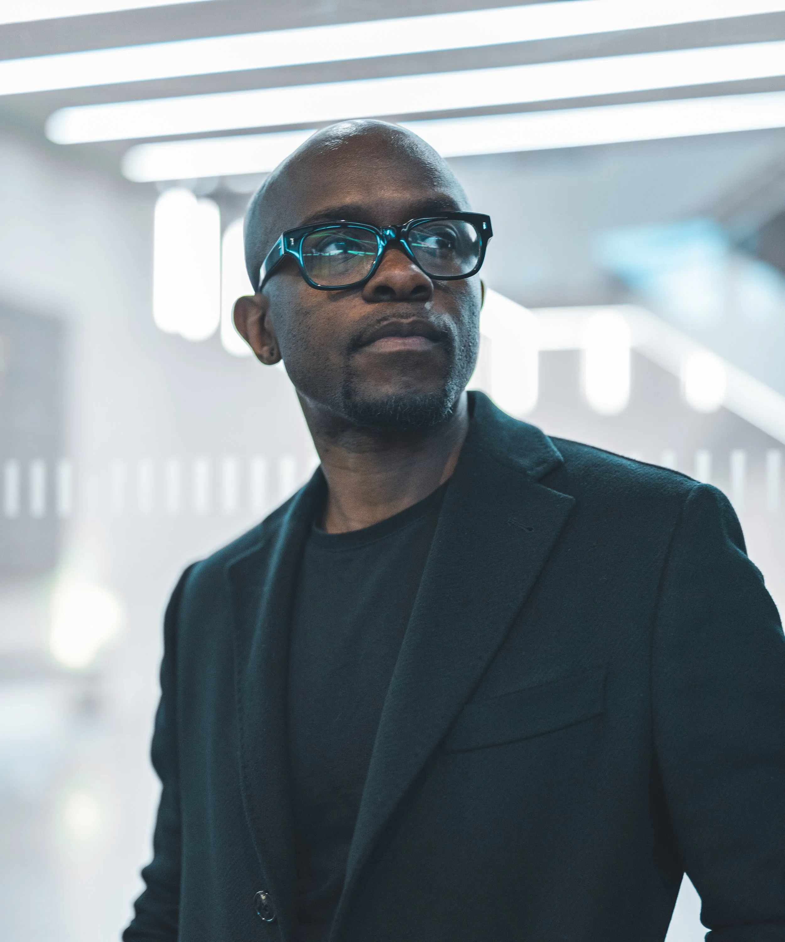 A man wearing glasses and a black blazer looks to the side in a modern indoor setting.