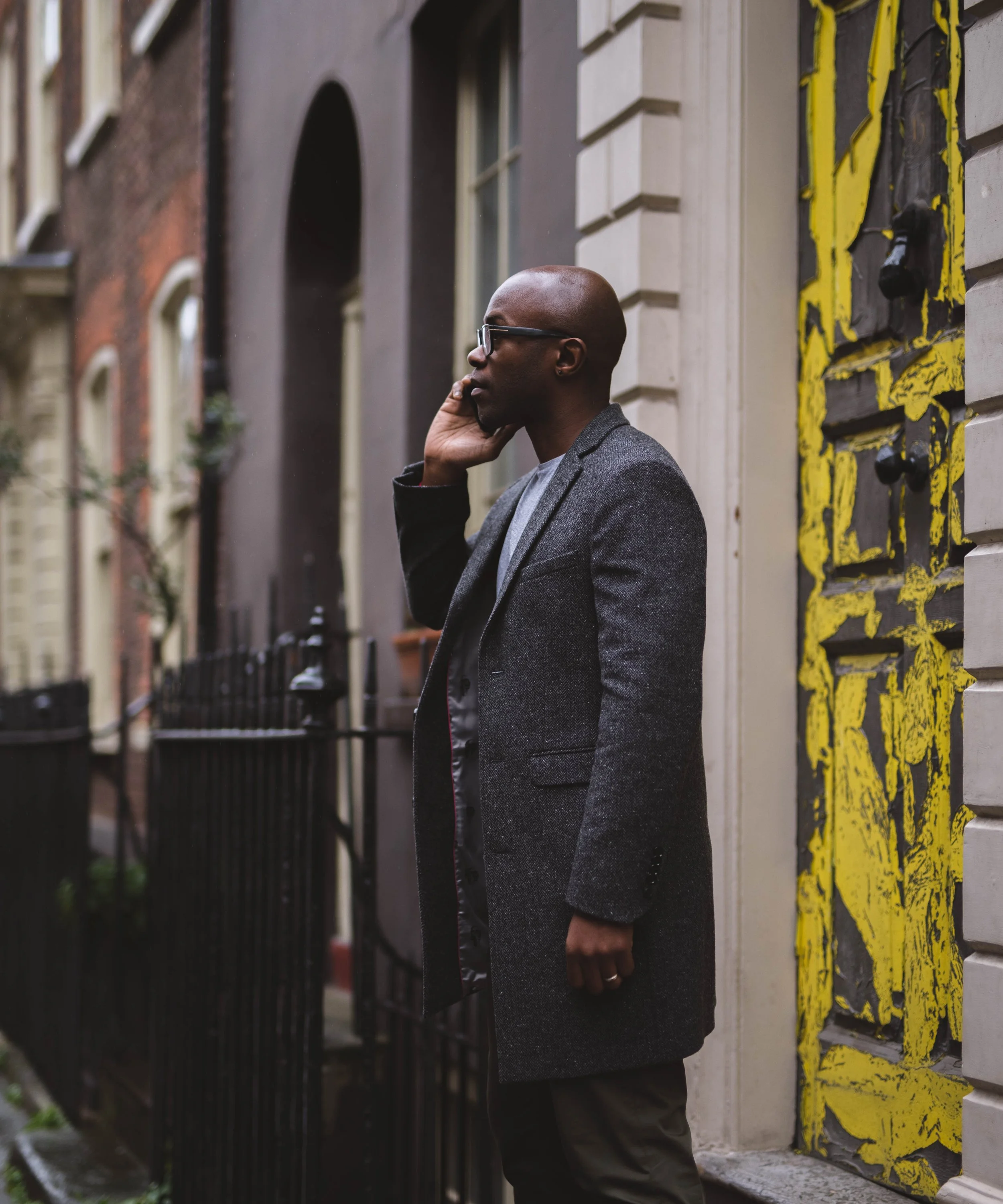 A man with glasses talking on his cellphone on a city sidewalk, standing next to a building with decorative yellow and black artwork.