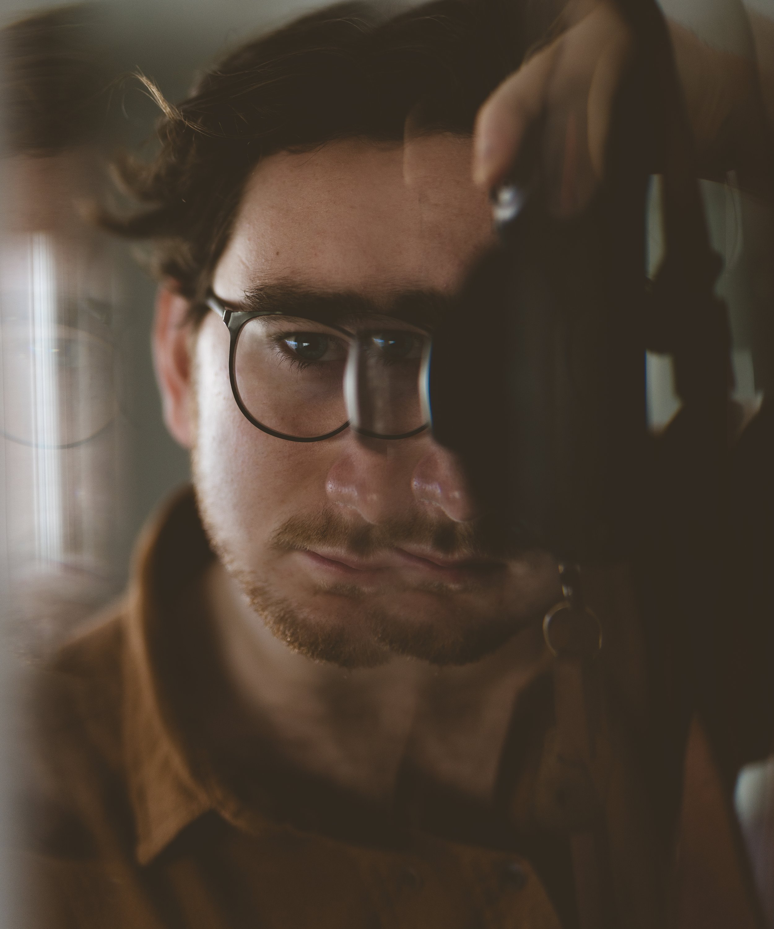 A man with glasses and a beard, looking thoughtfully through a camera lens, in a close-up portrait shot.