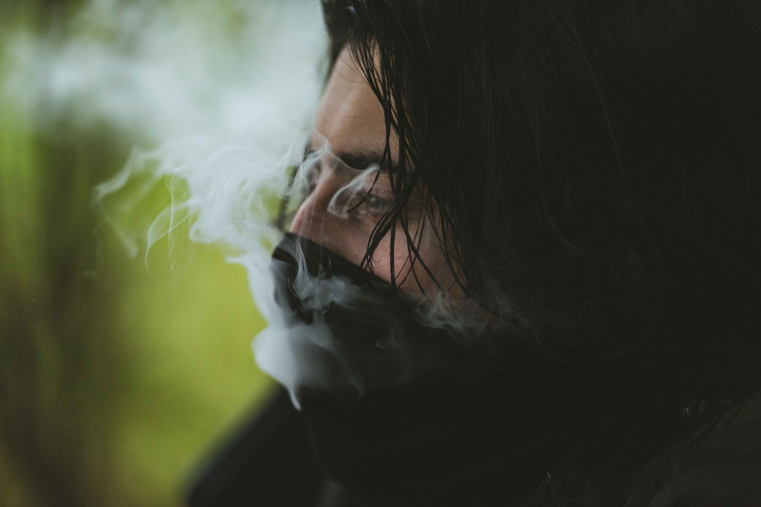 A person with black hair and glasses exhaling smoke outdoors with greenery blurred in the background.