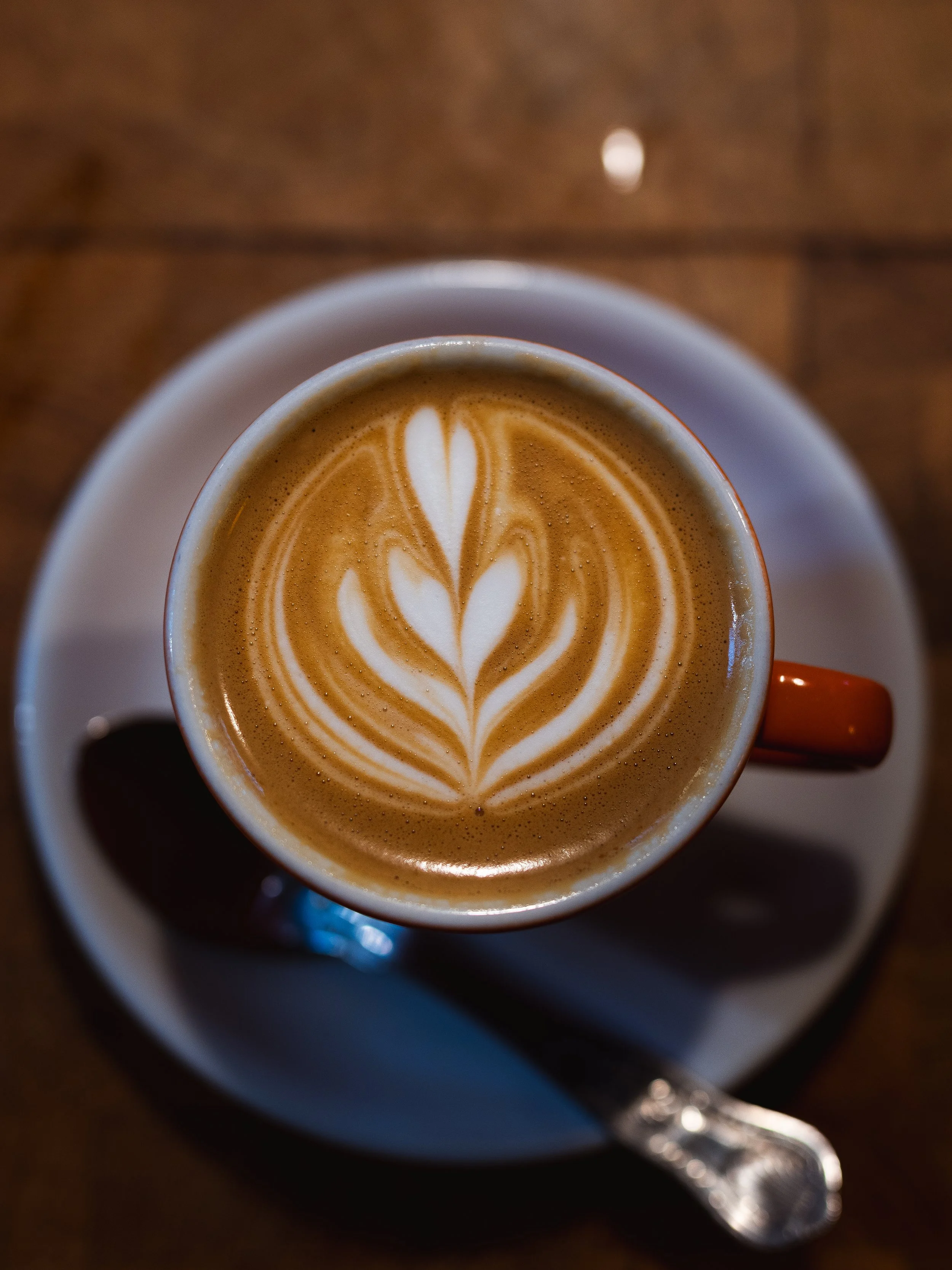 A top view of a cup of latte with latte art on top, placed on a saucer with a spoon, on a wooden table.