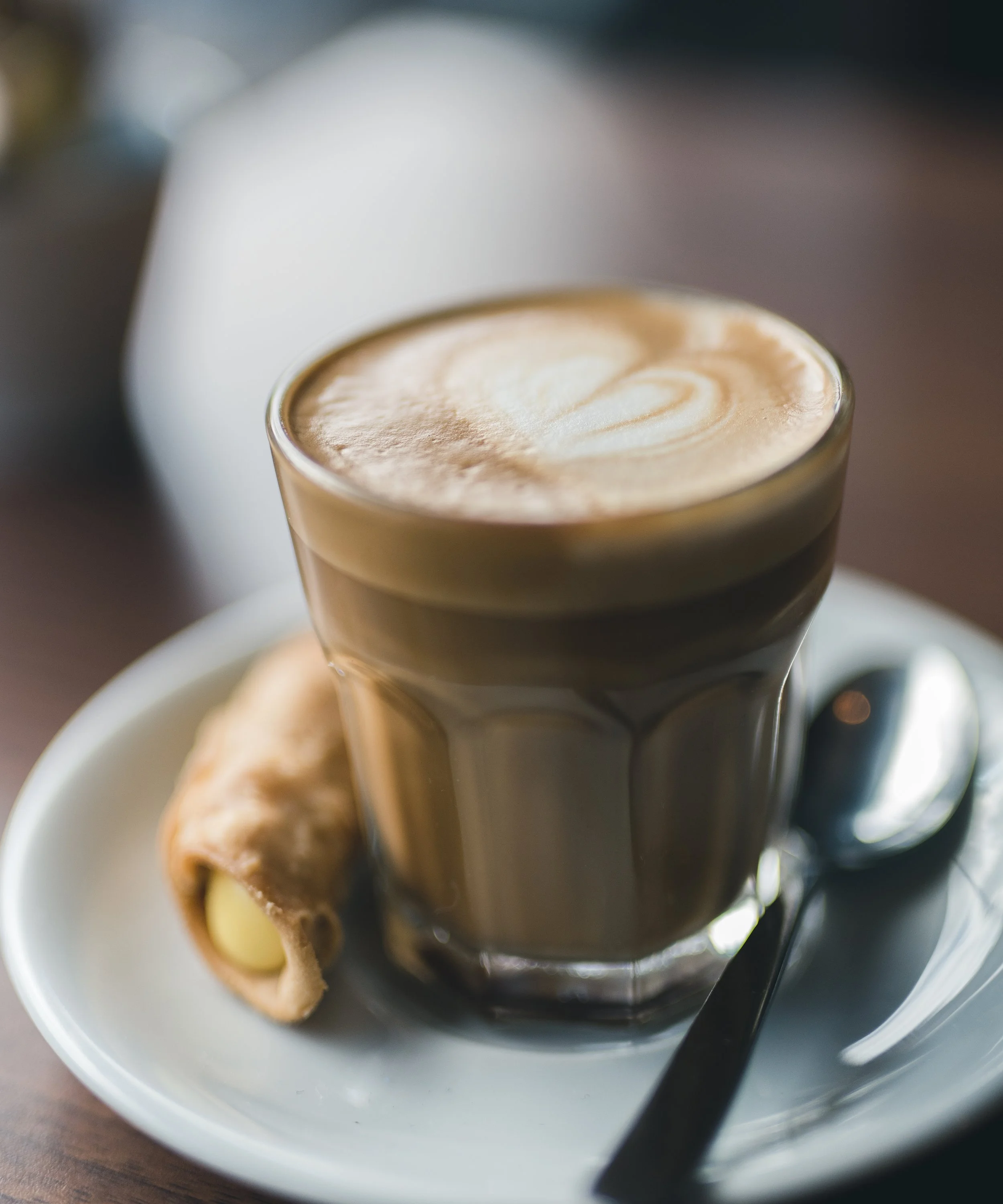 A glass of coffee with foam on top, served on a saucer with a teaspoon and a gingerbread cookie.