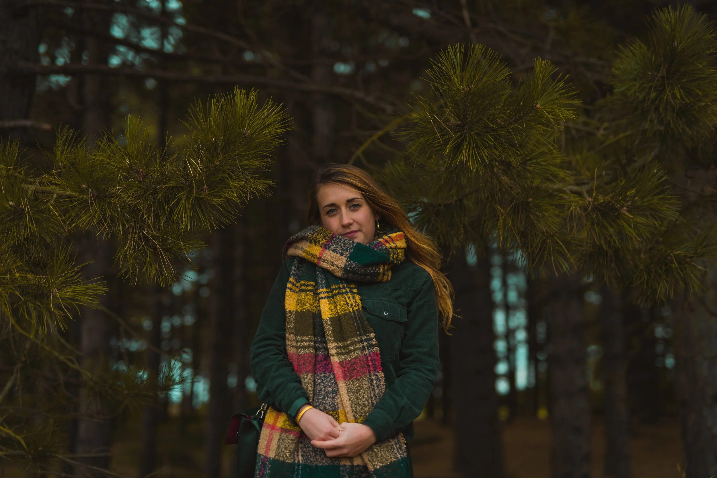 A young woman with long, light brown hair standing outdoors among pine trees, wearing a green jacket and a multicolored plaid scarf.