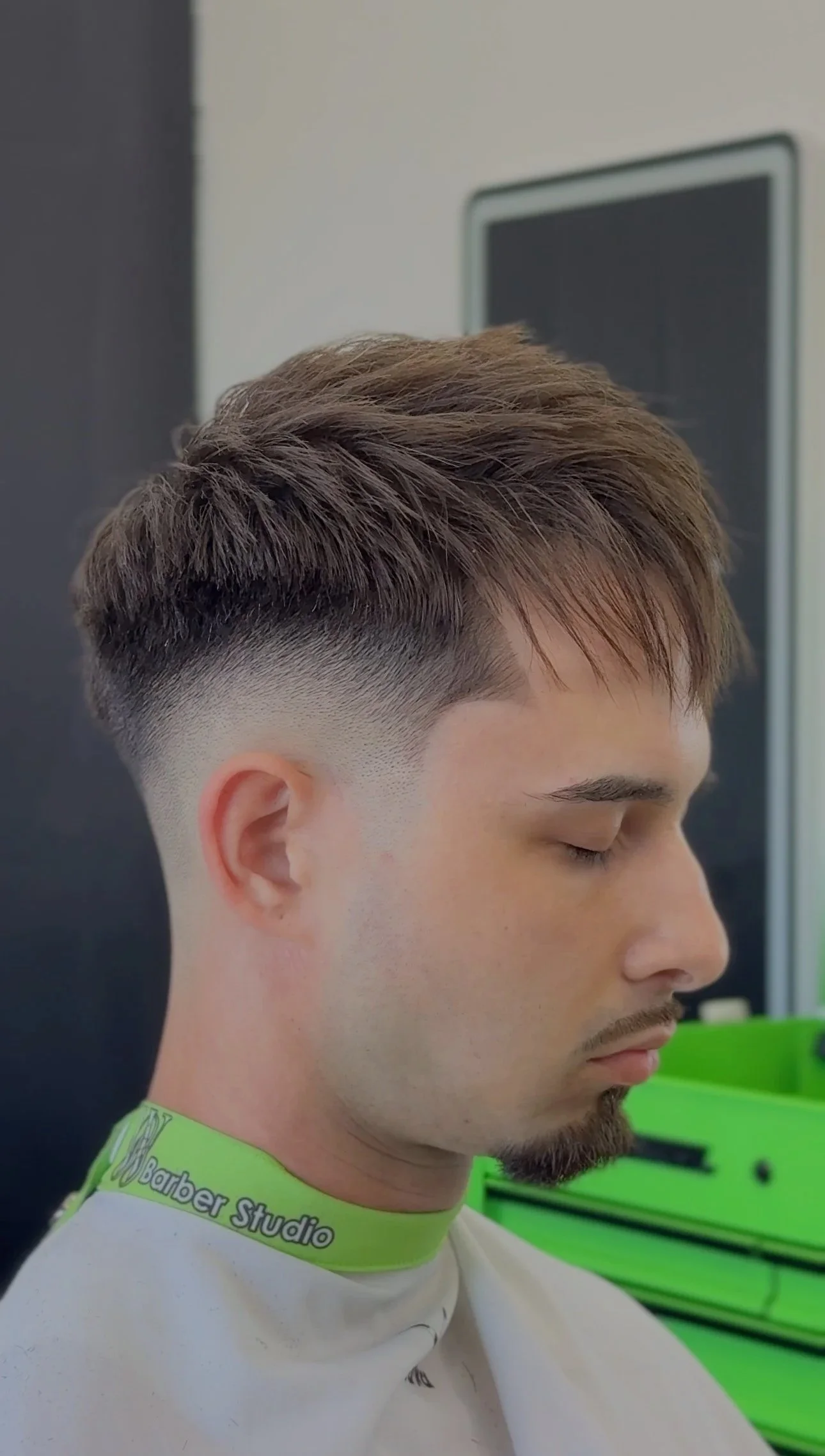 Hombre con corte de cabello moderna, con degradado en los lados y peinado hacia arriba, sentado en una silla en una barbería.