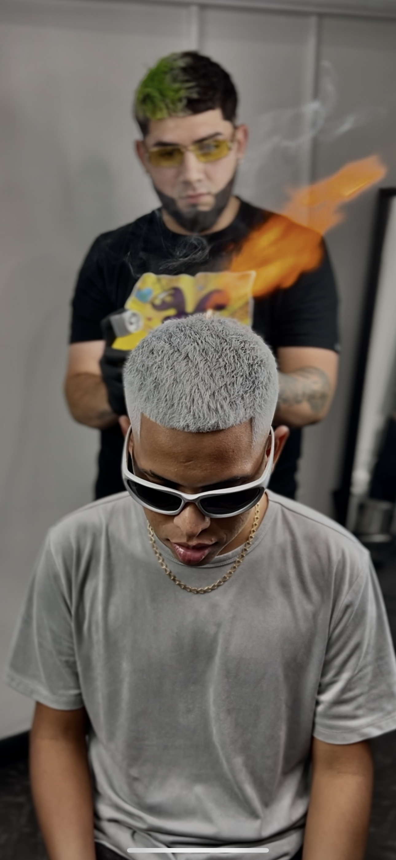 Un hombre con cabello rubio y corto, usando gafas negras, sentado con la cabeza inclinada hacia abajo, y otro hombre con cabello teñido de gris con verde, usando gafas amarillas, sosteniendo una fuente de fuego.