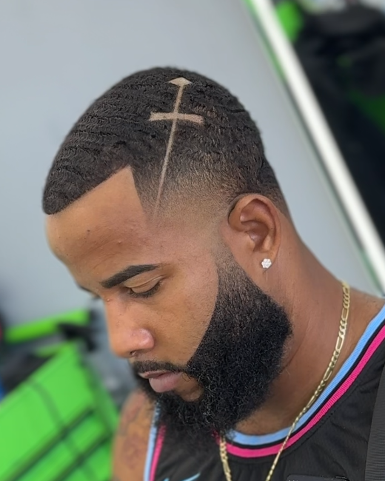 Hombre con peinado de corte afilado y diseño de cruz en el cabello, barba tupida, pendiente de diamante y cadena, looks de barbería.