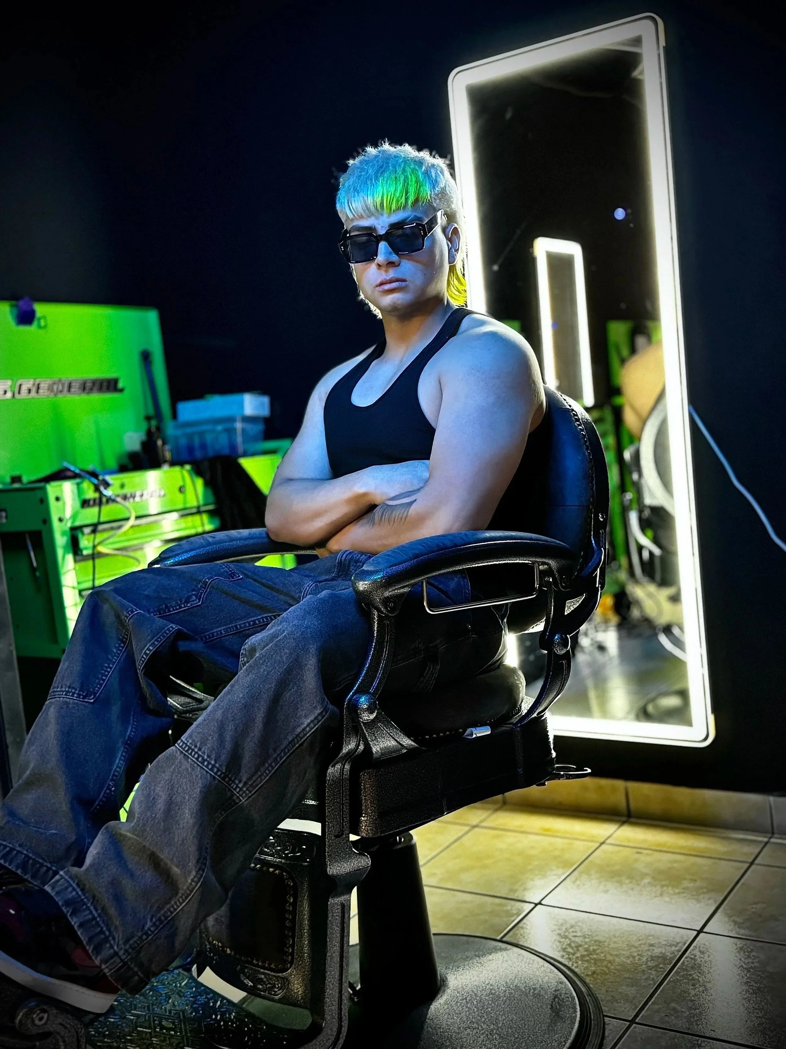 Persona con cabello multicolor, lentes oscuros y ropa deportiva, sentado en una silla en un entorno con espejos de luz y equipo en un salón moderno.