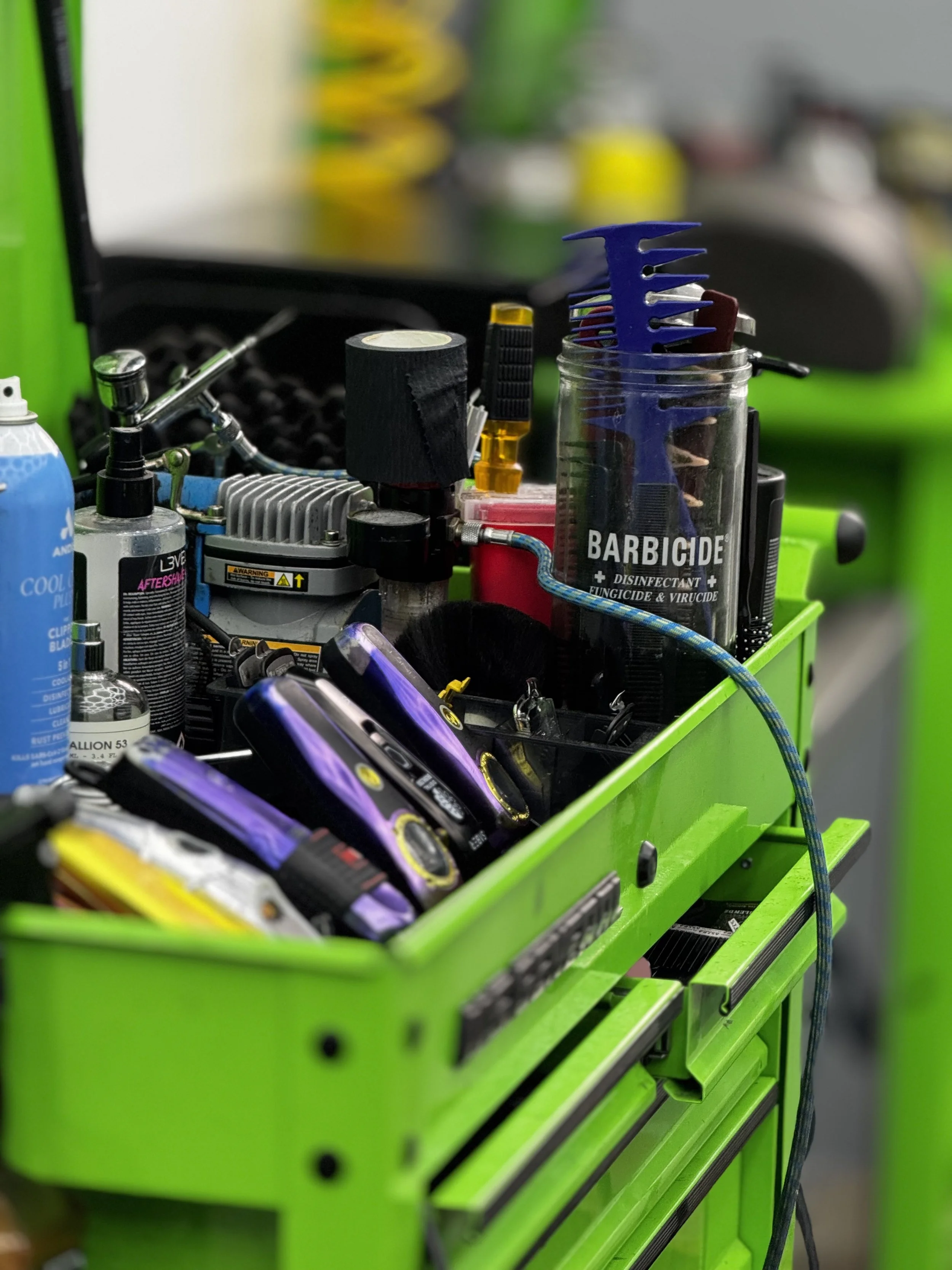 Cajón de herramientas verde con varios objetos, incluyendo una lata de desinfectante, varios detectores de mano, un recipiente con herramientas, un equipo de gasolina, y otros utensilios de mecánica o reparación.