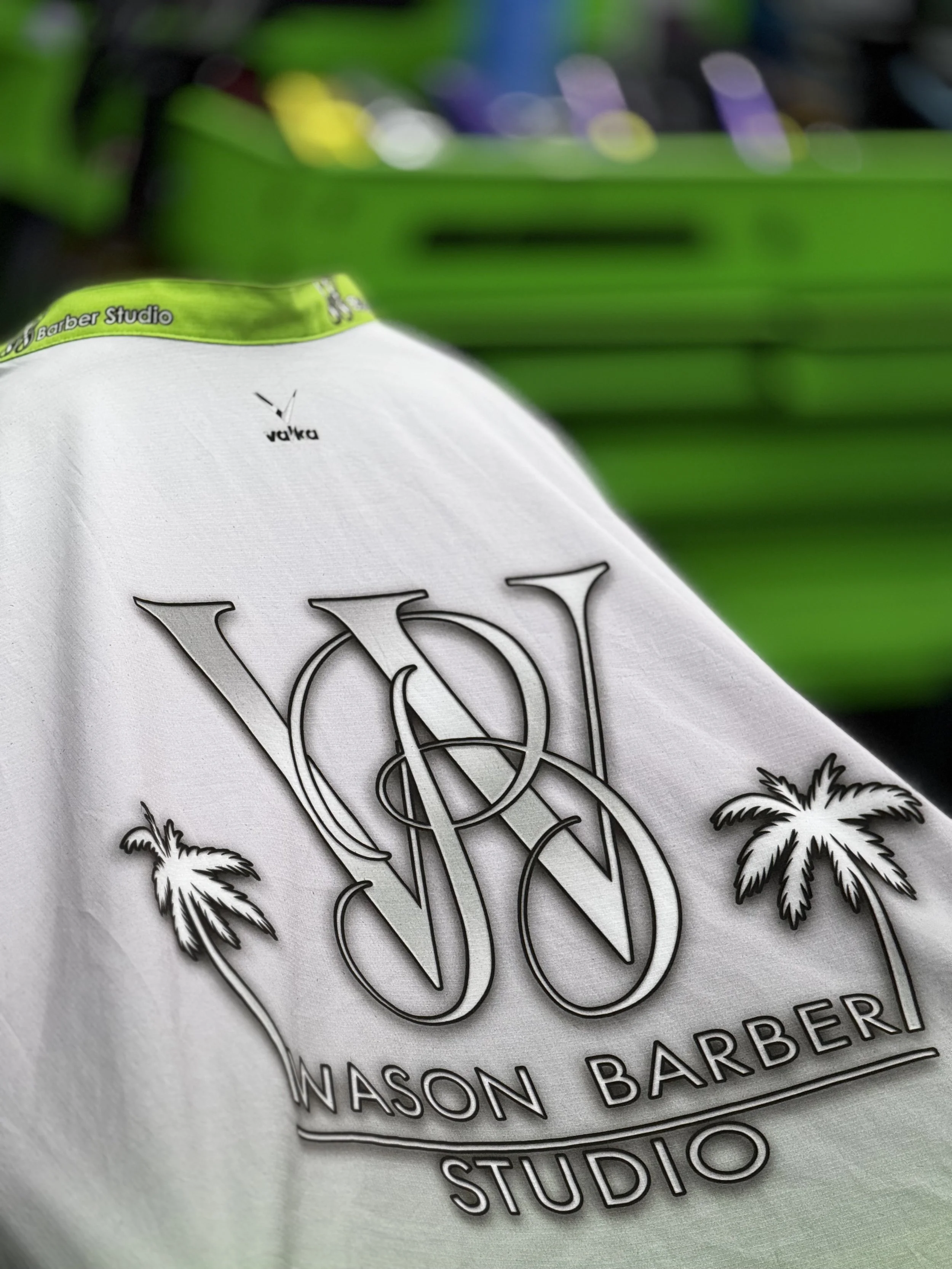 Camiseta blanca con el logotipo del estudio de barbería Wason Barber Studio y dibujos de palmeras, con fondo de mobiliario verde.