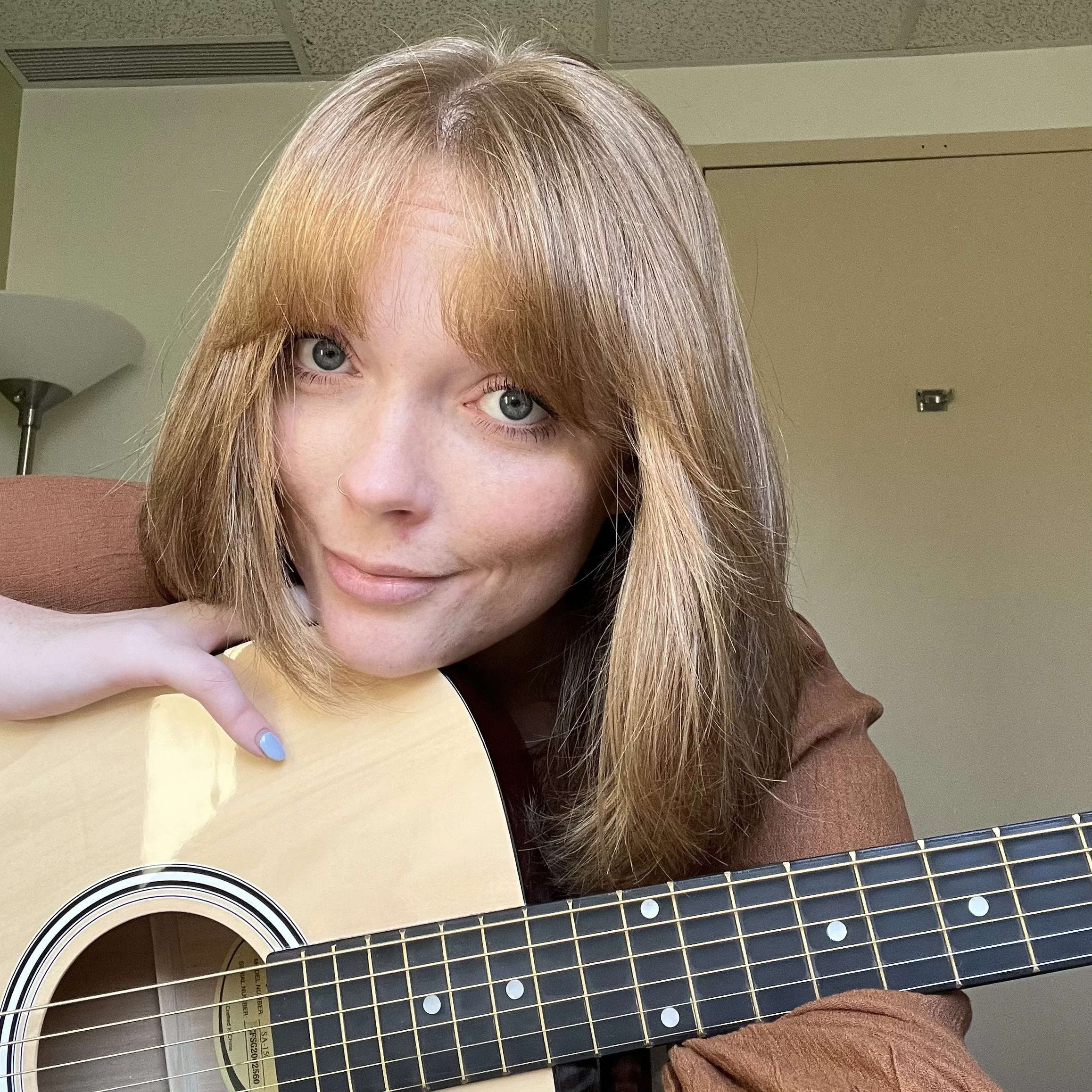 A woman with red hair, blue eyes, and a small nose ring playing an acoustic guitar.