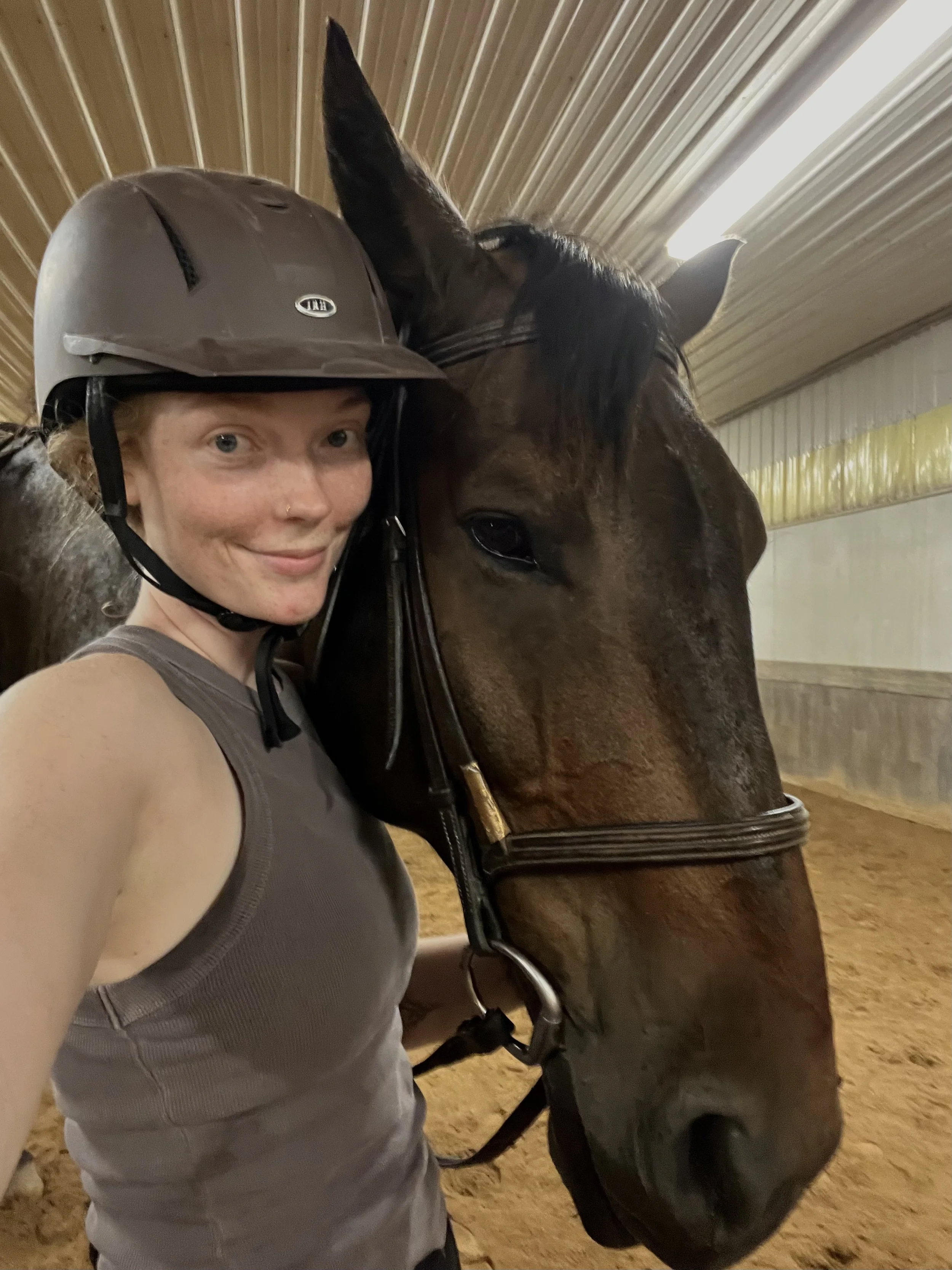 A woman in a helmet and sleeveless shirt taking a selfie with a horse inside a stable.