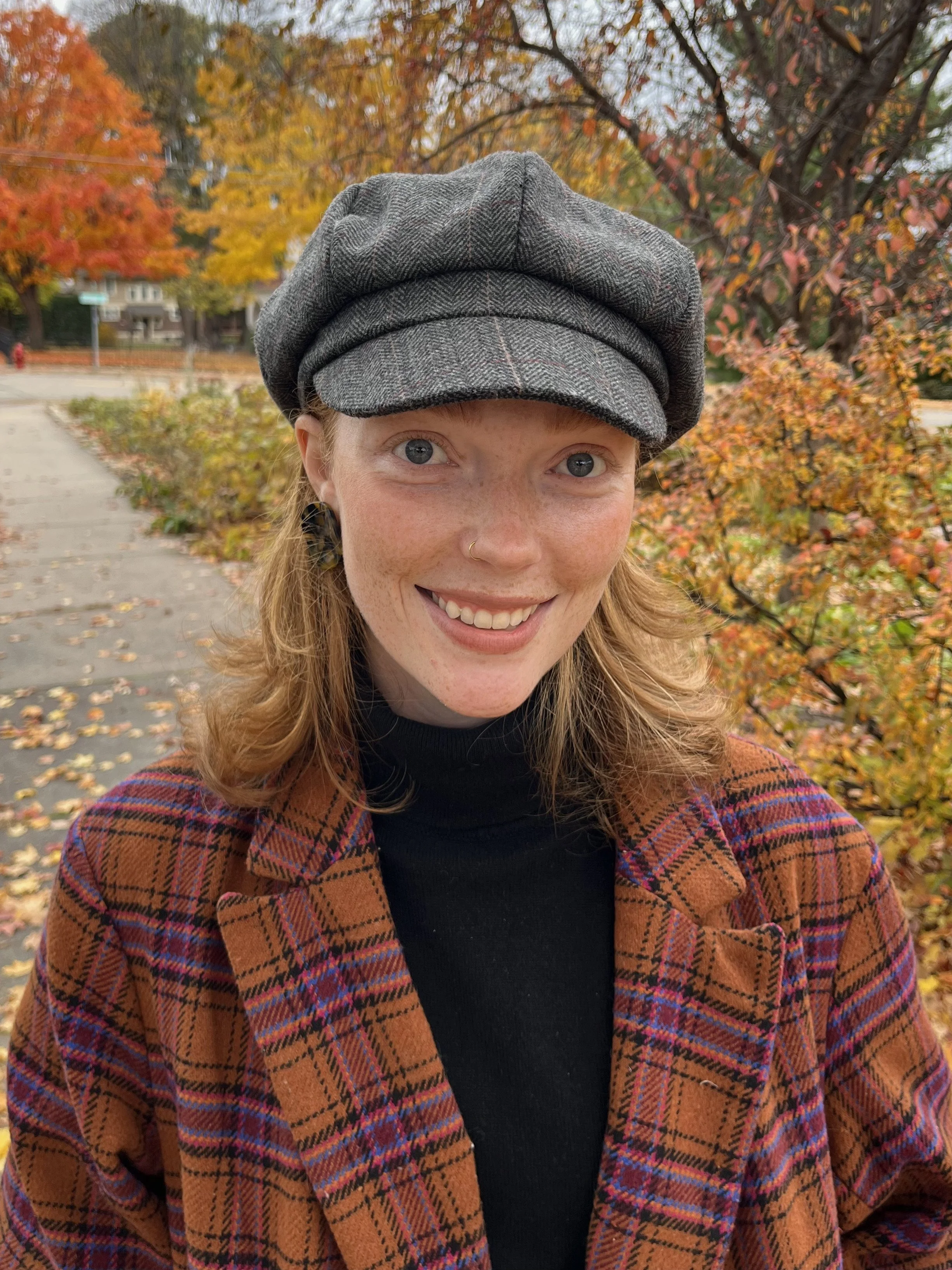 A smiling woman with red hair, wearing a gray plaid newsboy cap, a black turtleneck, and a brown plaid jacket standing outdoors on an autumn day with colorful fall foliage in the background.