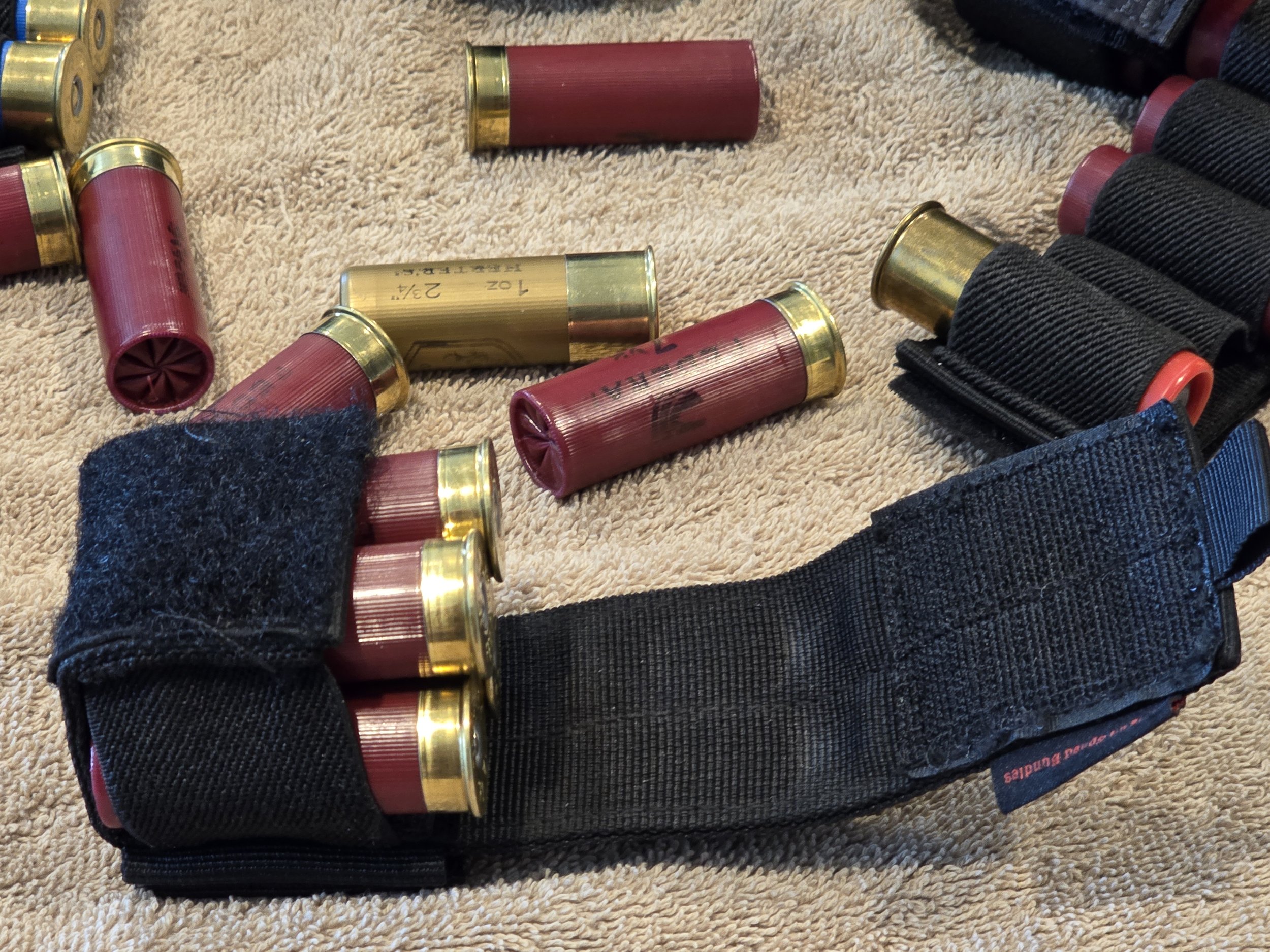 A black tactical gun magazine holder with some shotgun shells inside, lying on a beige carpet with more shotgun shells spread out nearby.