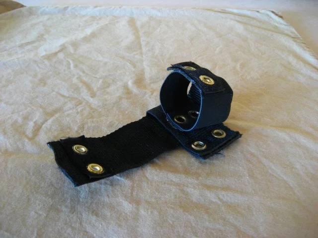 A black fabric armband with multiple metal eyelets, partially unrolled, on a crinkled white surface.