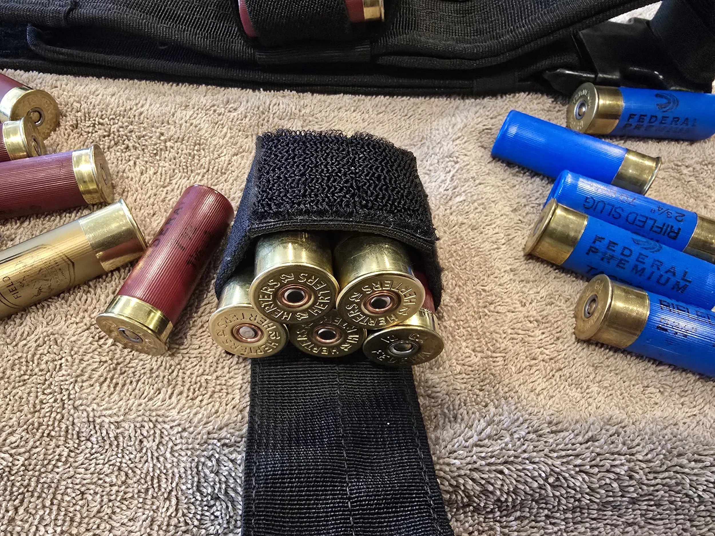 Shotgun shells in red and blue colors scattered on a beige towel, with some inside a black holder and others outside. The shells have brass bases, some with visible markings, and the scene suggests reloading or disposal of ammunition.