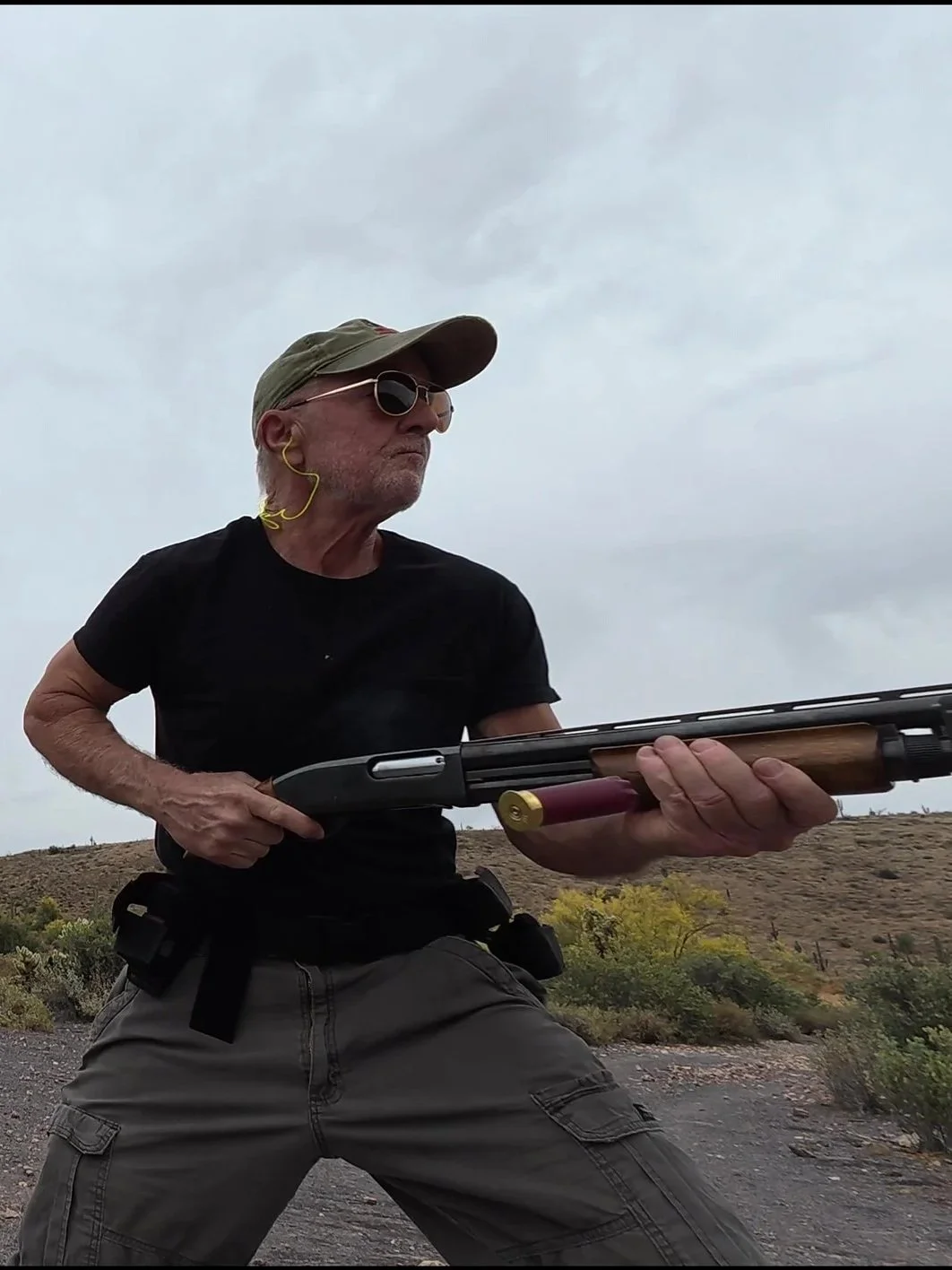 An elderly man outdoors holding a rifle, wearing sunglasses, a cap, and ear protection, with desert landscape in the background.