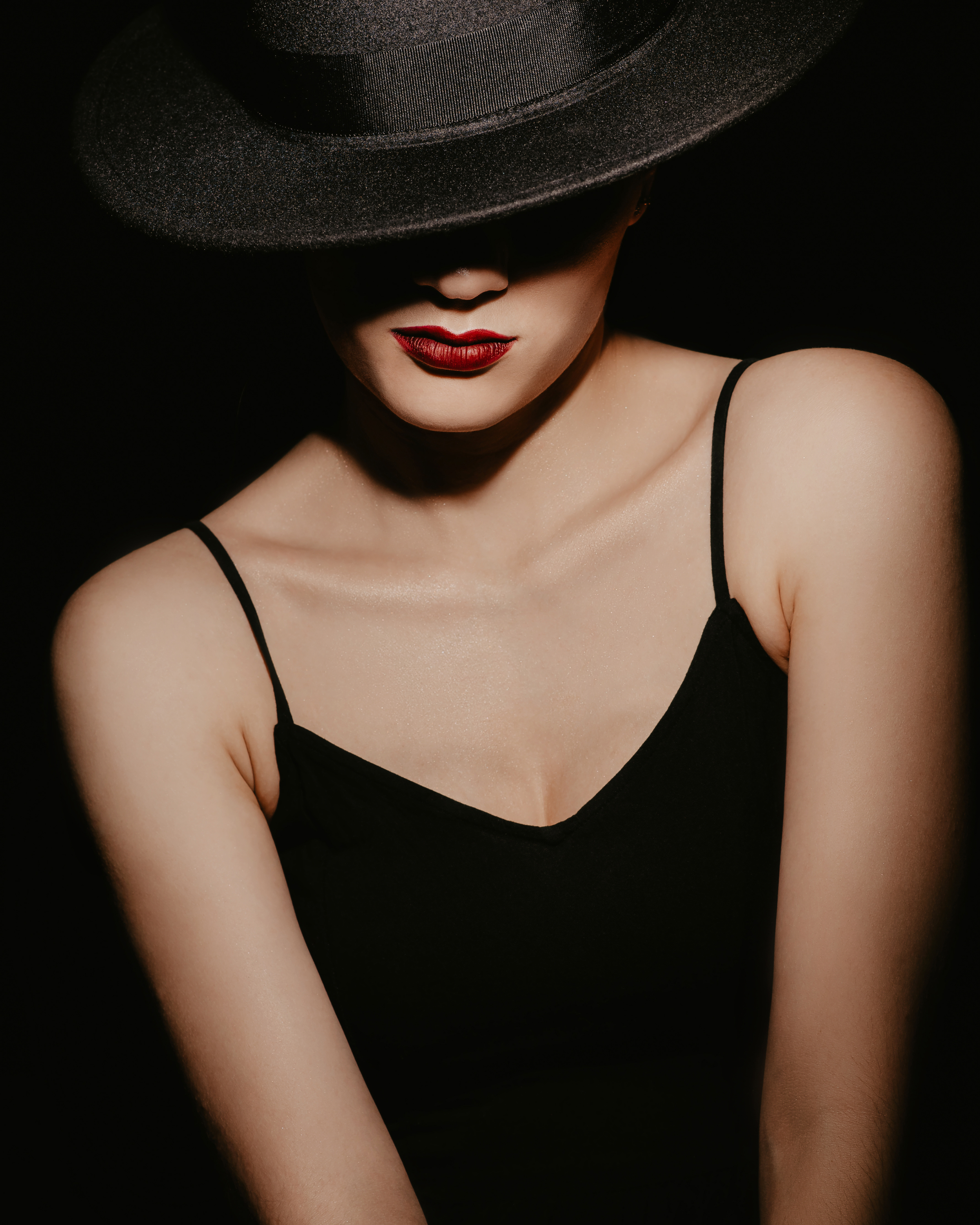A woman wearing a large black hat, with her face partially obscured, dressed in a black spaghetti strap top, with red lipstick and smooth skin. Femme fatale.