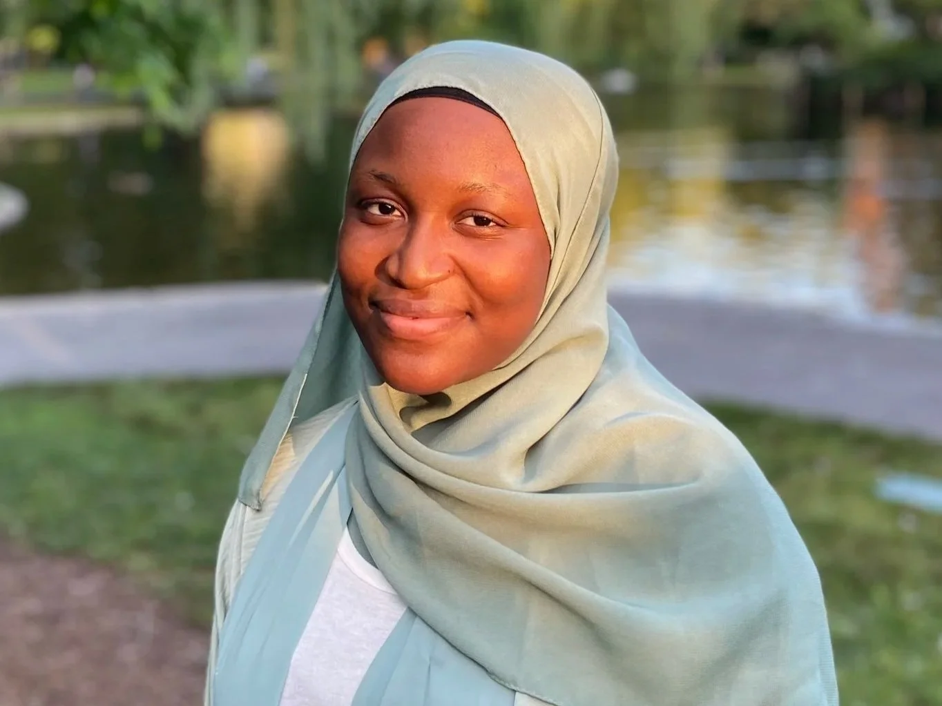 A woman wearing a light-colored hijab smiling outdoors near a body of water with a park in the background.