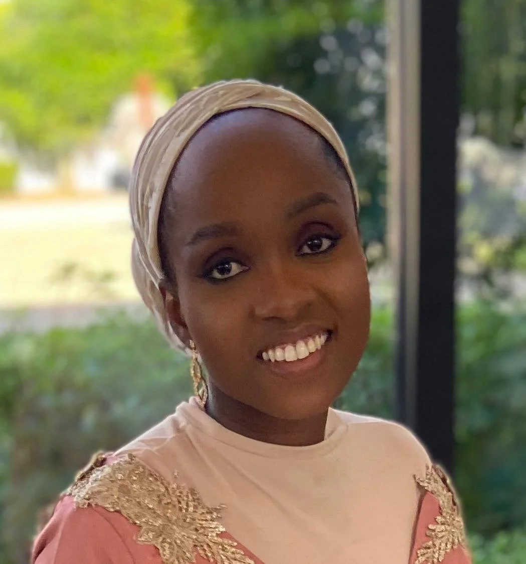 A woman with a beige headscarf and a pink dress with gold embroidery, smiling inside near a window with outdoor greenery in the background.