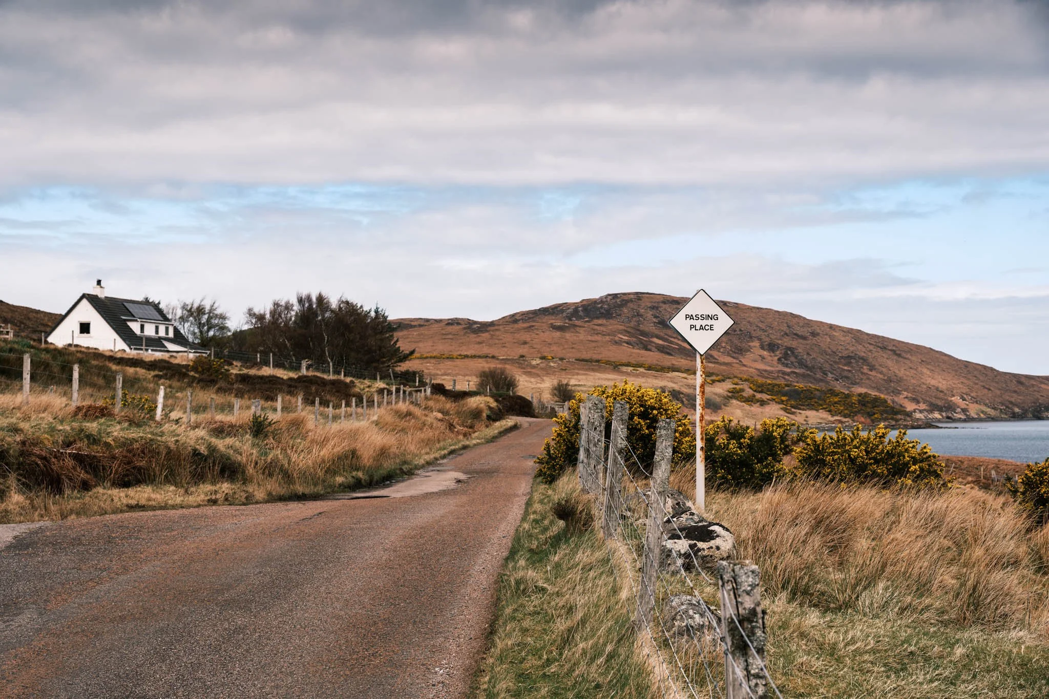 Highland - Scotland - Une route de terre bordée d'une clôture en bois, avec un panneau de signalisation 'Passing Place' devant un paysage vallonné, un Loch, un ciel nuageux et une maison blanche dans la végétation environnante.