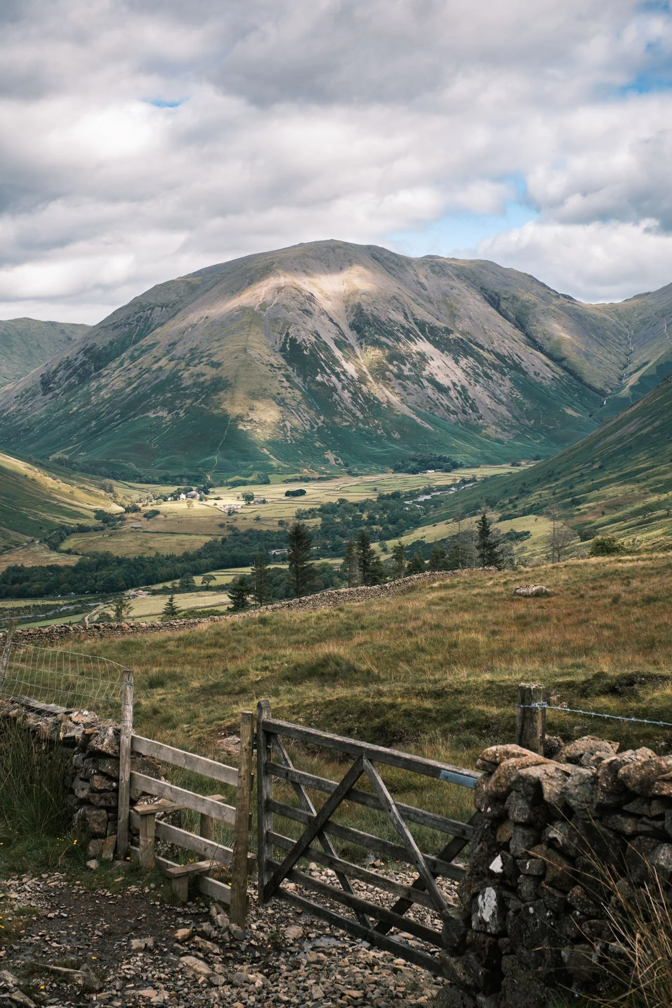 Lake District - Un paysage de montagne avec des collines verdoyantes et un ciel nuageux, avec une clôture en bois et un portail en premier plan.  Reportage outdoor, mise en valeur du territoire et tourisme durable par Guillaume Donsimoni.