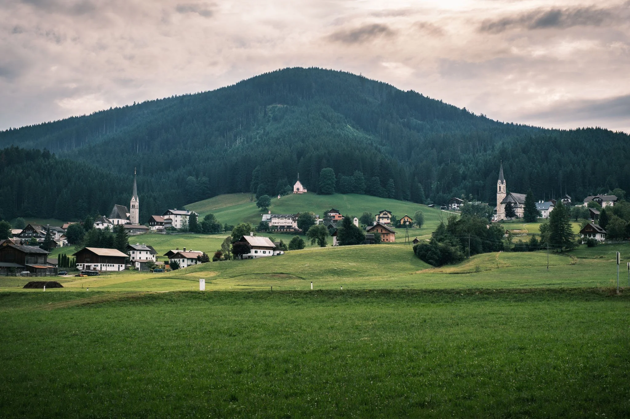 Austria - Un village paisible avec des maisons et deux églises, entouré de champs verts et situé près d'une grande forêt, avec une montagne en arrière-plan, sous un ciel nuageux. Reportage outdoor, mise en valeur du territoire et tourisme durable par
