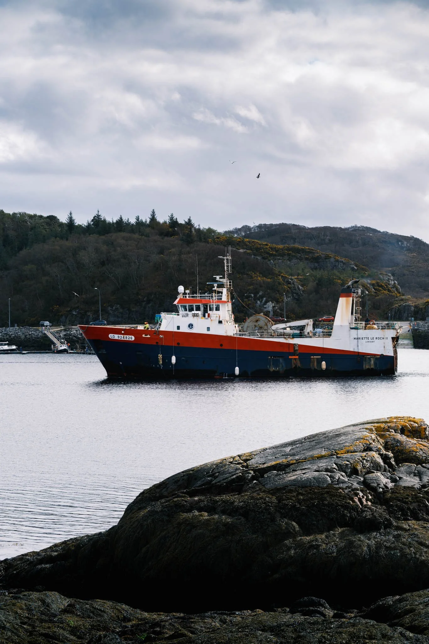 Highland - Scotland - Un bateau de pêche rouge et blanc amarré dans une baie avec la mer calme, des collines boisées en arrière-plan et un ciel nuageux.