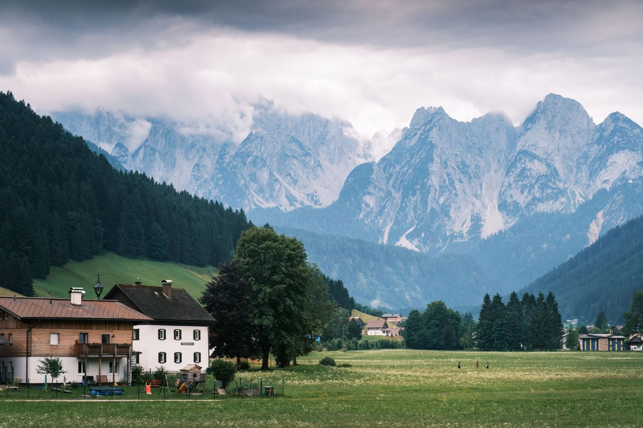Tyrol - Paysage de montagne avec des maisons, des arbres et un champ herbeux devant une chaîne de montagnes rocheuses sous un ciel nuageux. Reportage outdoor, mise en valeur du territoire et tourisme durable par Guillaume Donsimoni