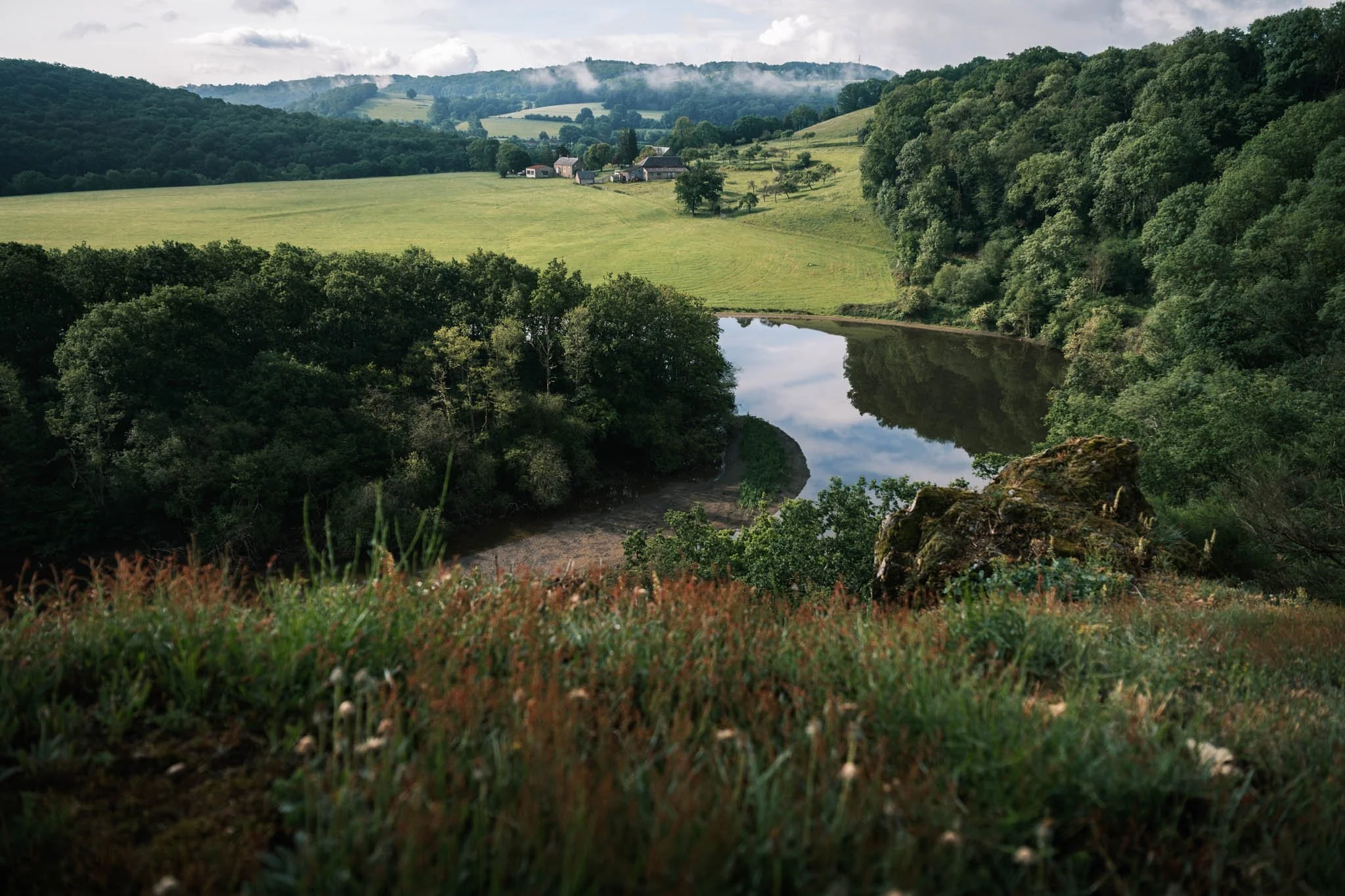 Suisse Normande - Paysage de campagne avec un ruisseau, des collines verdoyantes, des arbres et un petit village en arrière-plan, sous un ciel partiellement nuageux.