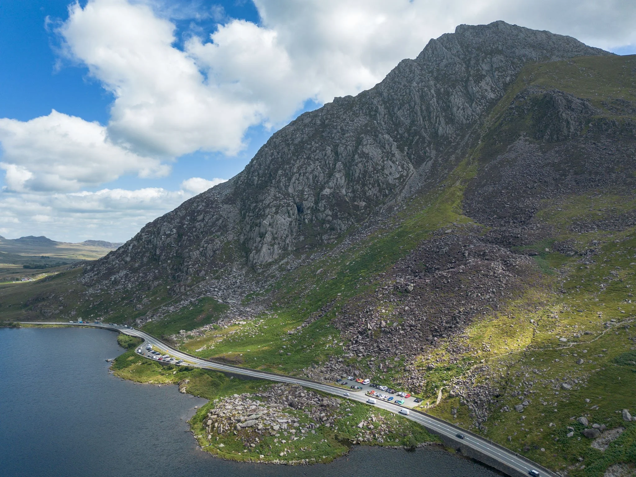 Wales - A road runs alongside a lake surrounded by rocky mountains under a partially cloudy sky. Snowdonia. Outdoor reportage, regional promotion, and sustainable tourism by Guillaume Donsimoni.