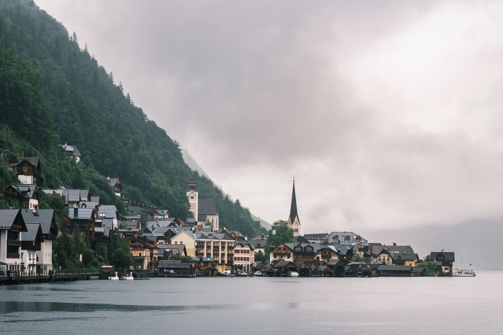 Austria - Village d'Hallstatt au bord d'un lac avec des maisons, une église avec un clocher pointu, et des montagnes verdoyantes en arrière-plan sous un ciel nuageux. Reportage outdoor, mise en valeur du territoire et tourisme durable par Guillaume D
