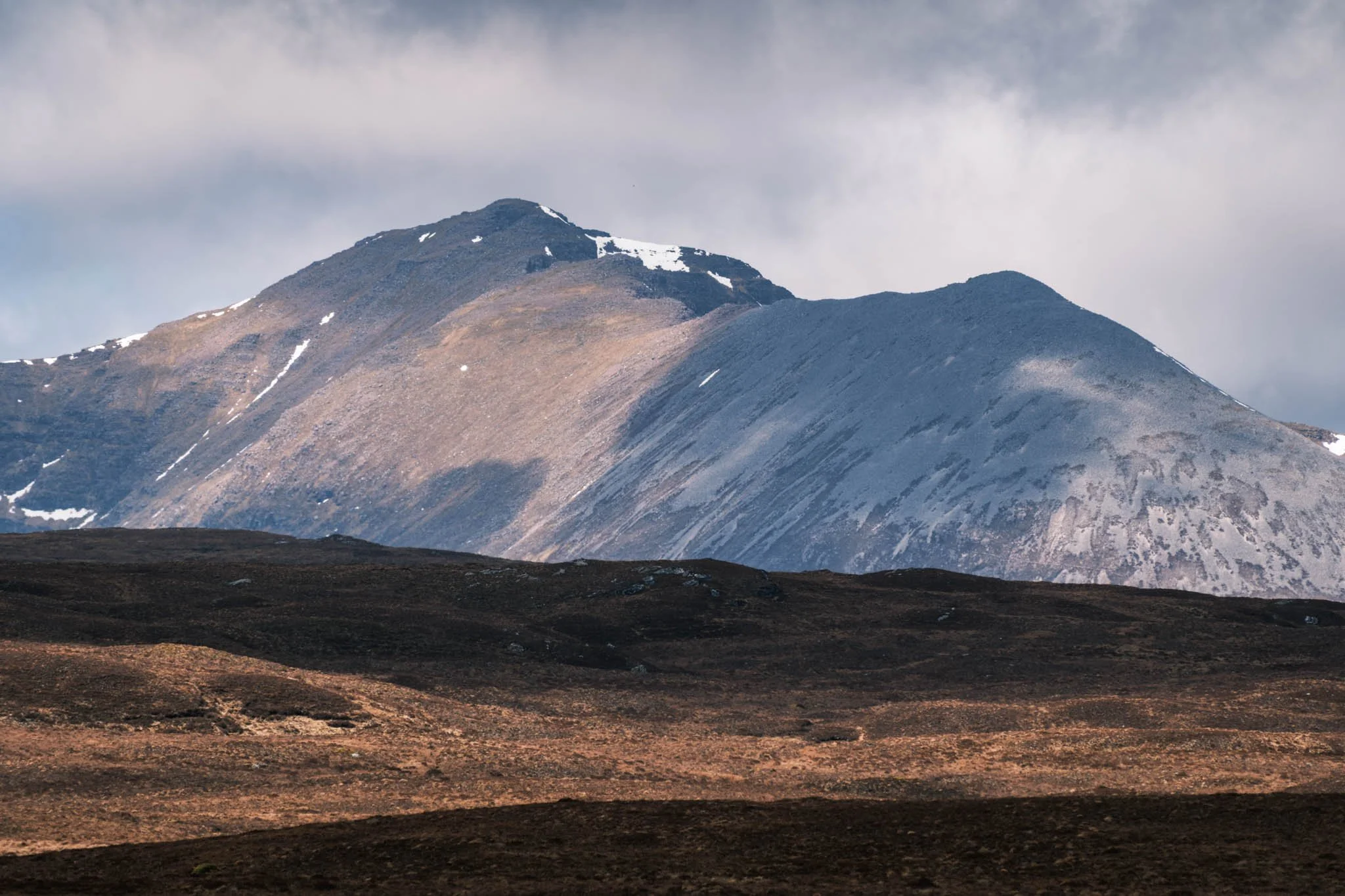 Highland - Scotland - Une montagne avec des restes de neige sur ses sommets, sous un ciel nuageux, avec un terrain aride en premier plan.
