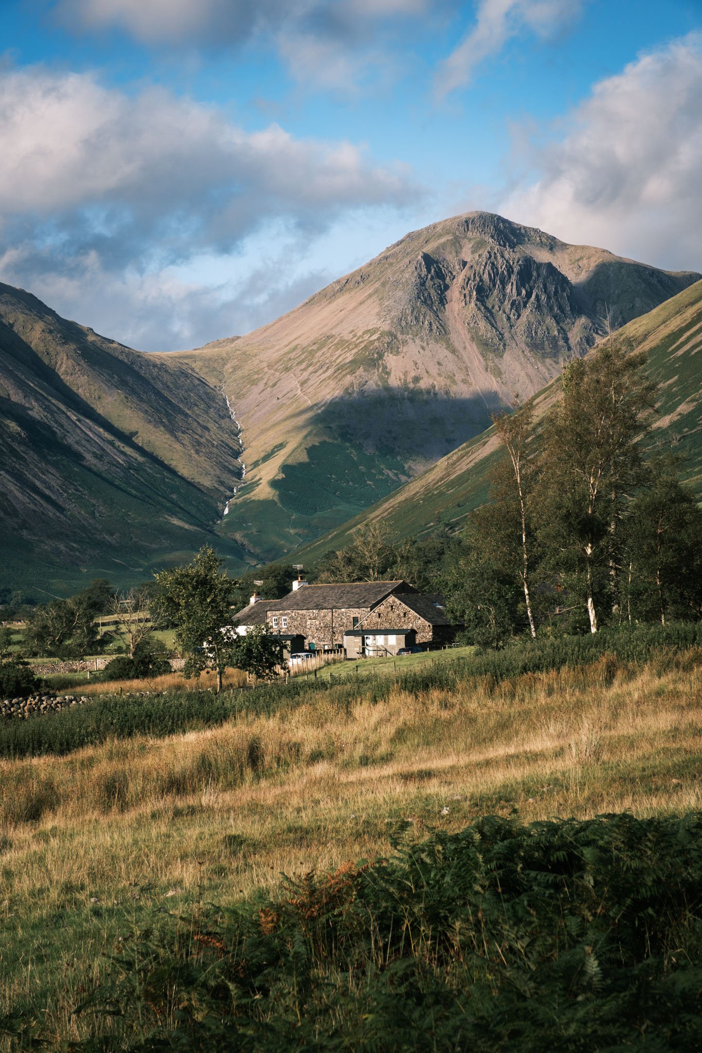Lake District - Paysage de montagne avec une maison en pierre, végétation verdoyante et ciel partiellement nuageux.  Reportage outdoor, mise en valeur du territoire et tourisme durable par Guillaume Donsimoni.