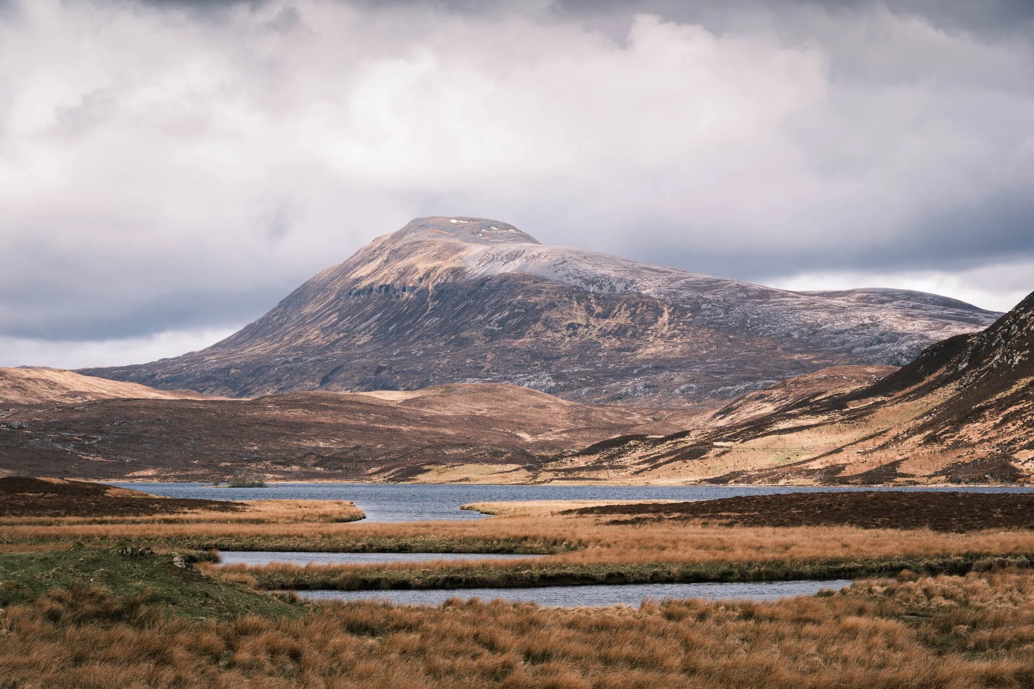 Highland - Scotland - Montagne avec sommet enneigé, terrain vallonné environnant, Loch au premier plan, ciel nuageux.