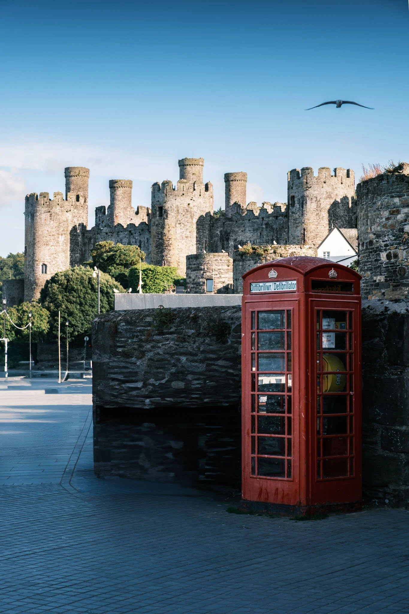 Wales - A red telephone booth in front of a medieval stone castle with towers, under a clear blue sky. Conwy Castle. Outdoor reportage, regional promotion, and sustainable tourism by Guillaume Donsimoni.