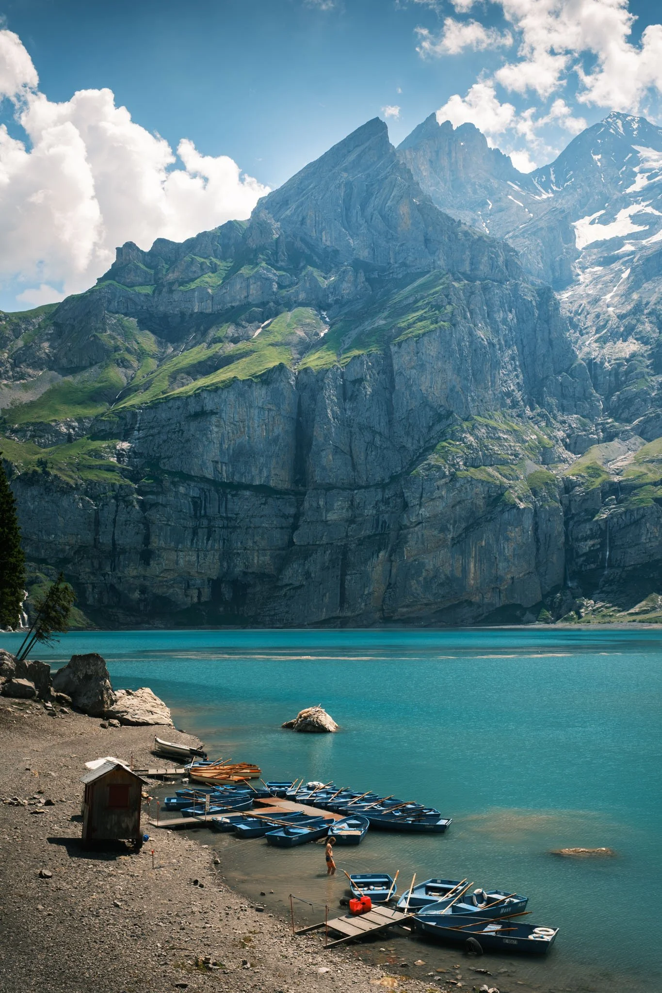 Oeschinensee. Lac de montagne avec des bateaux amarrés à la rive, entouré de falaises rocheuses et de montagnes enneigées au loin, sous un ciel partiellement nuageux. Reportage outdoor, mise en valeur du territoire et tourisme durable par Guillaume D