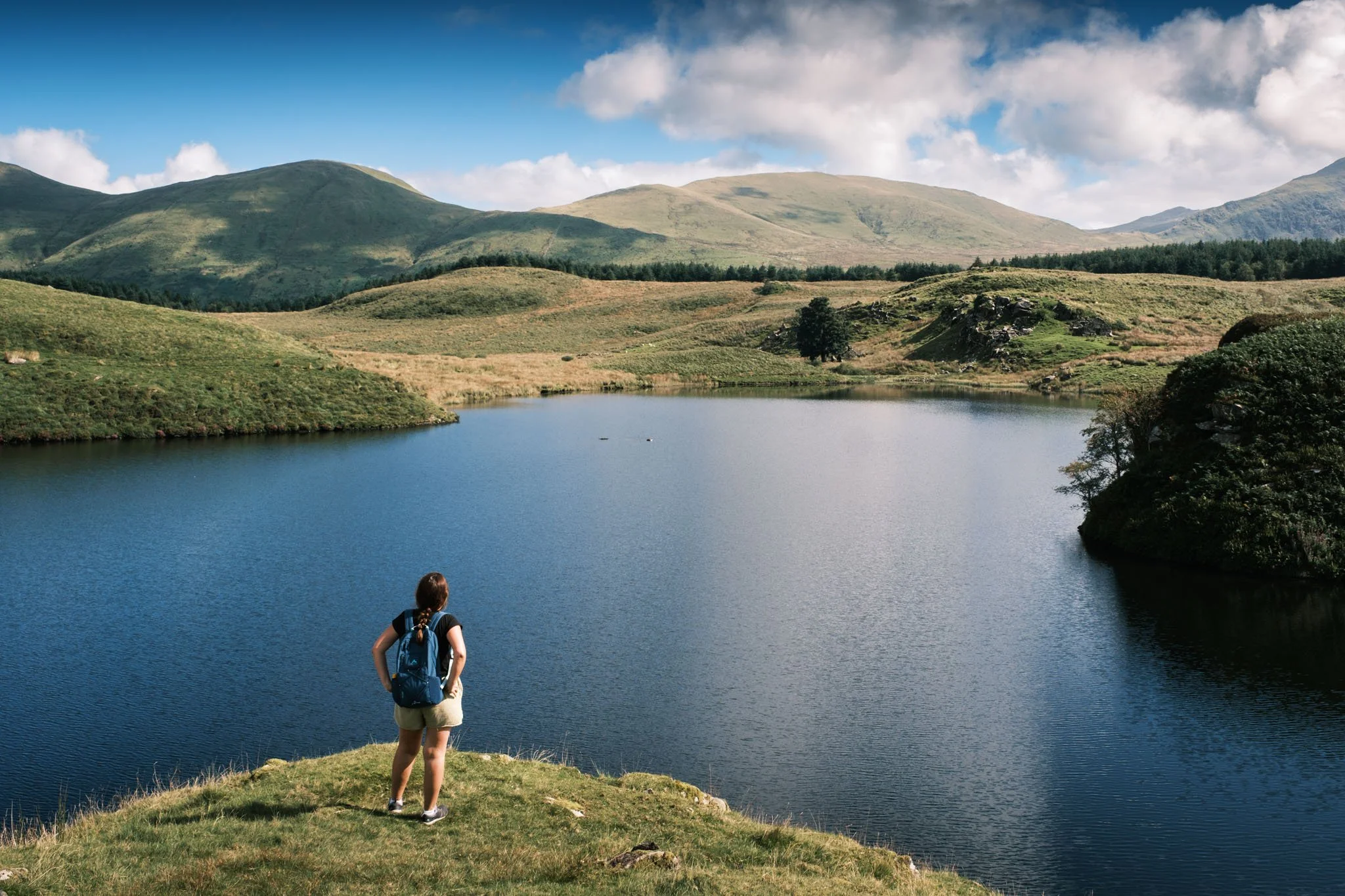 Wales - A woman with a backpack stands on a bank, looking at a lake surrounded by green hills under a partially cloudy sky. Outdoor reportage, regional promotion, and sustainable tourism by Guillaume Donsimoni.
